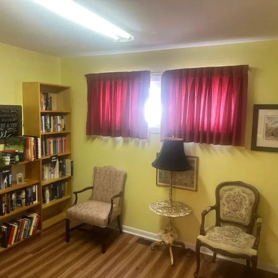 Small yellow room with bookshelves, two chairs, a side table, and red curtains over a bright window.