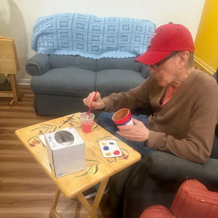 Person in a red cap sits at a small table, eating from a bowl beside a drink in a cozy living room.