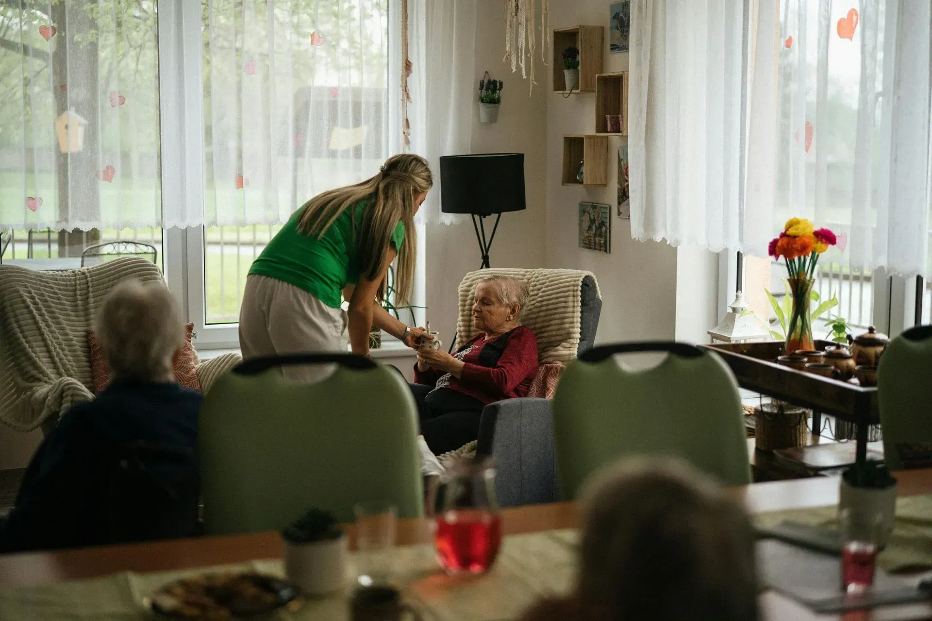 Caregiver leans toward seated resident in a bright lounge with large windows and tables.