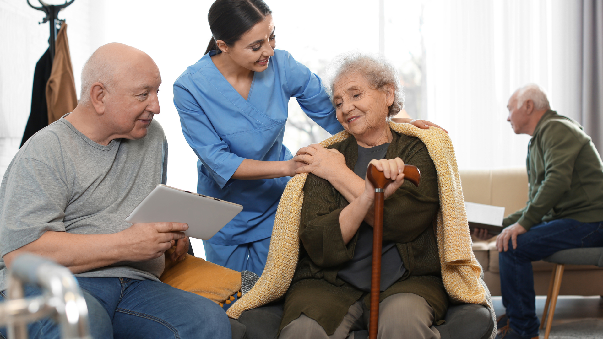 Caregiver in blue helping a smiling senior woman with a cane while two others sit nearby in a care home
