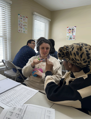 Three students collaborating at a classroom table, one holding a small craft item, with worksheets in the foreground