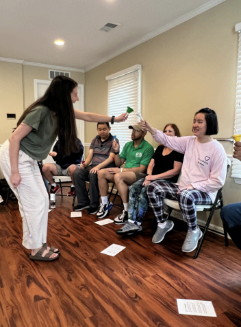 Woman in white pants interacts with seated people in a beige room during a group activity.