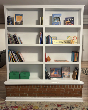 White bookshelf with books, games, and green bins against a brick wall backdrop