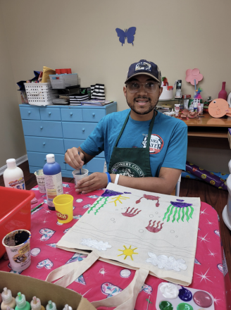 Person painting a white tote bag at a craft table with colorful paint supplies.