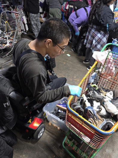 Person sorting items in a crowded outdoor market, wearing gloves and a backpack with a cart beside them