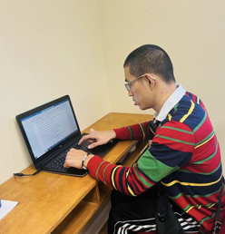 Person typing on a laptop at a wooden desk in a beige room, wearing a red striped sweater.
