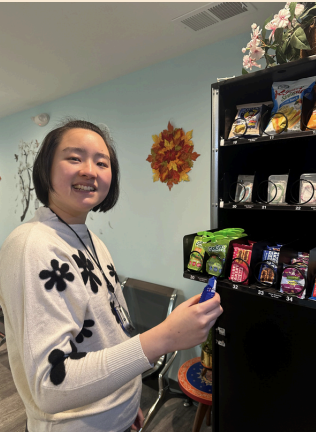 Smiling person in a room, holding a blue can beside a shelf of snacks and drinks.