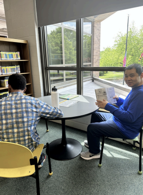 Two people sit at a table by large windows in a library, reading papers.