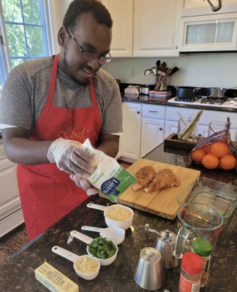 Person in a red apron seasoning food in a bright kitchen, with ingredients and utensils on the counter
