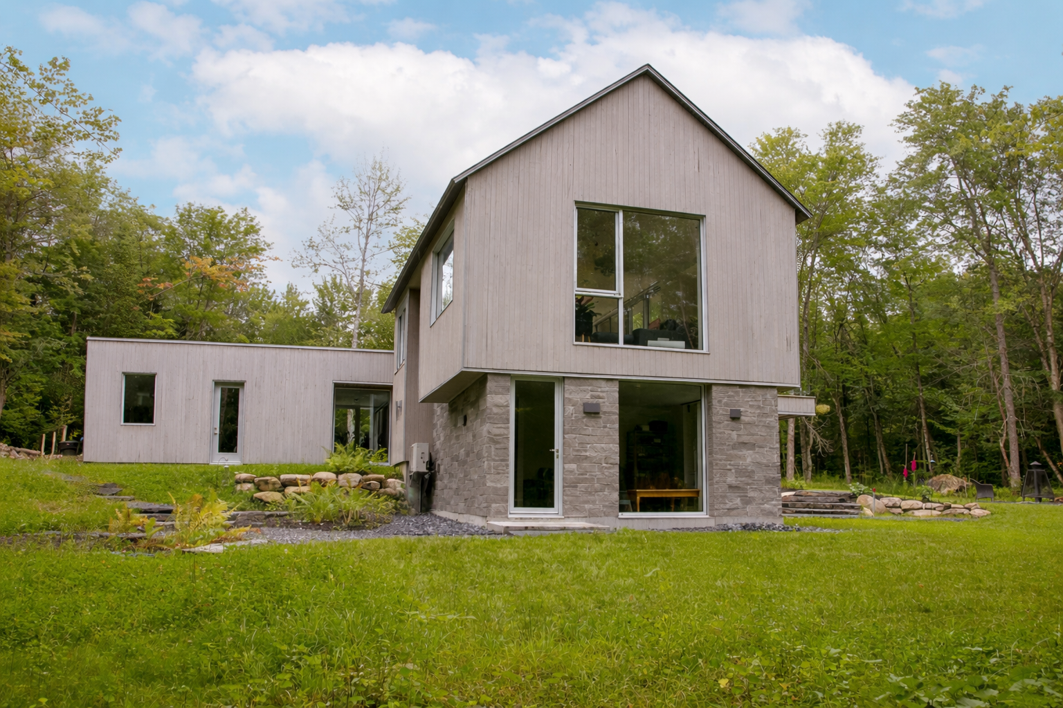 Une maison moderne à deux étages, avec un bardage en bois clair et une maçonnerie en pierre, nichée dans un cadre boisé luxuriant et verdoyant.