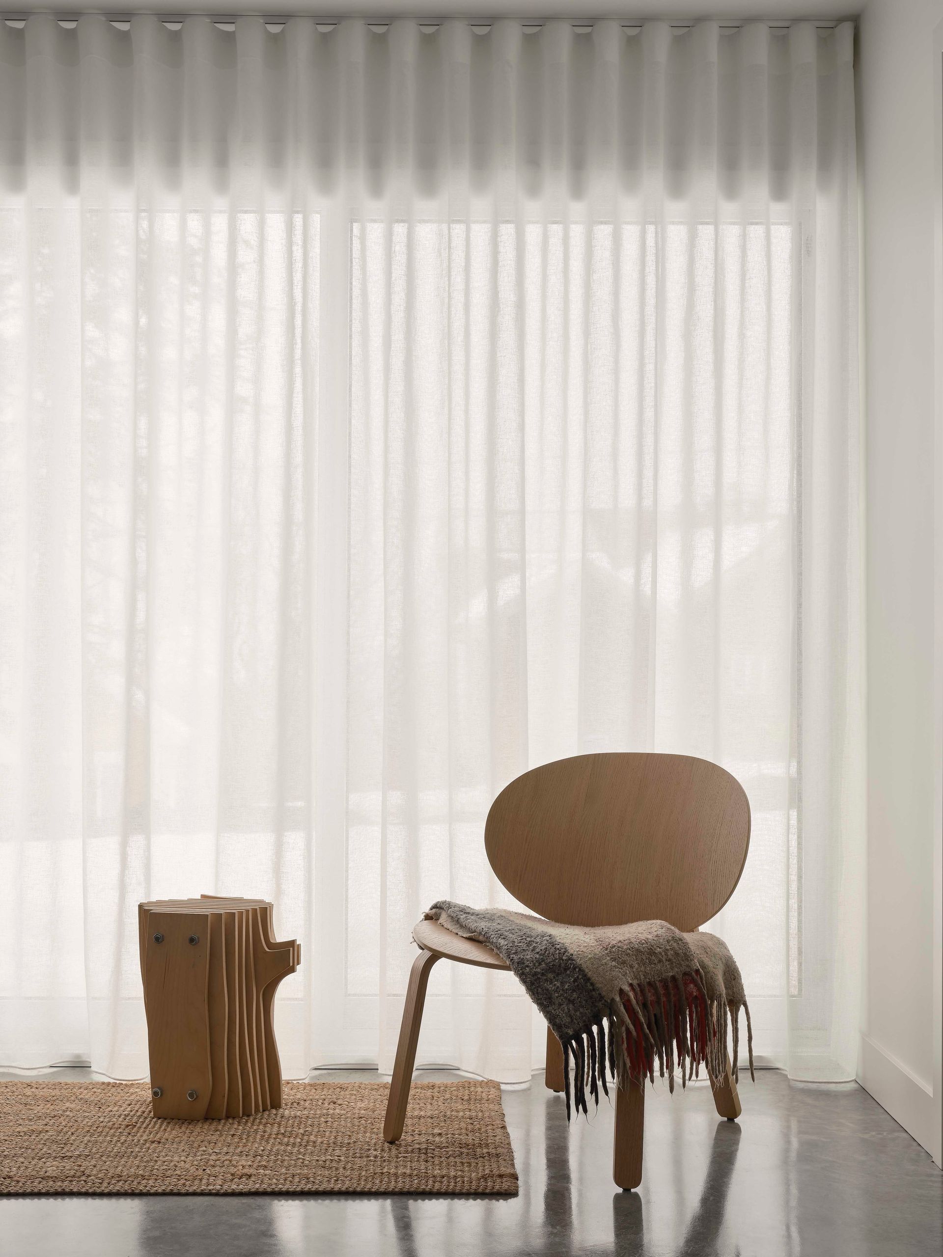 Une chaise en bois recouverte d'un plaid textur&eacute; et un petit tabouret en bois sont pos&eacute;s sur un tapis devant des rideaux blancs transparents allant du sol au plafond.