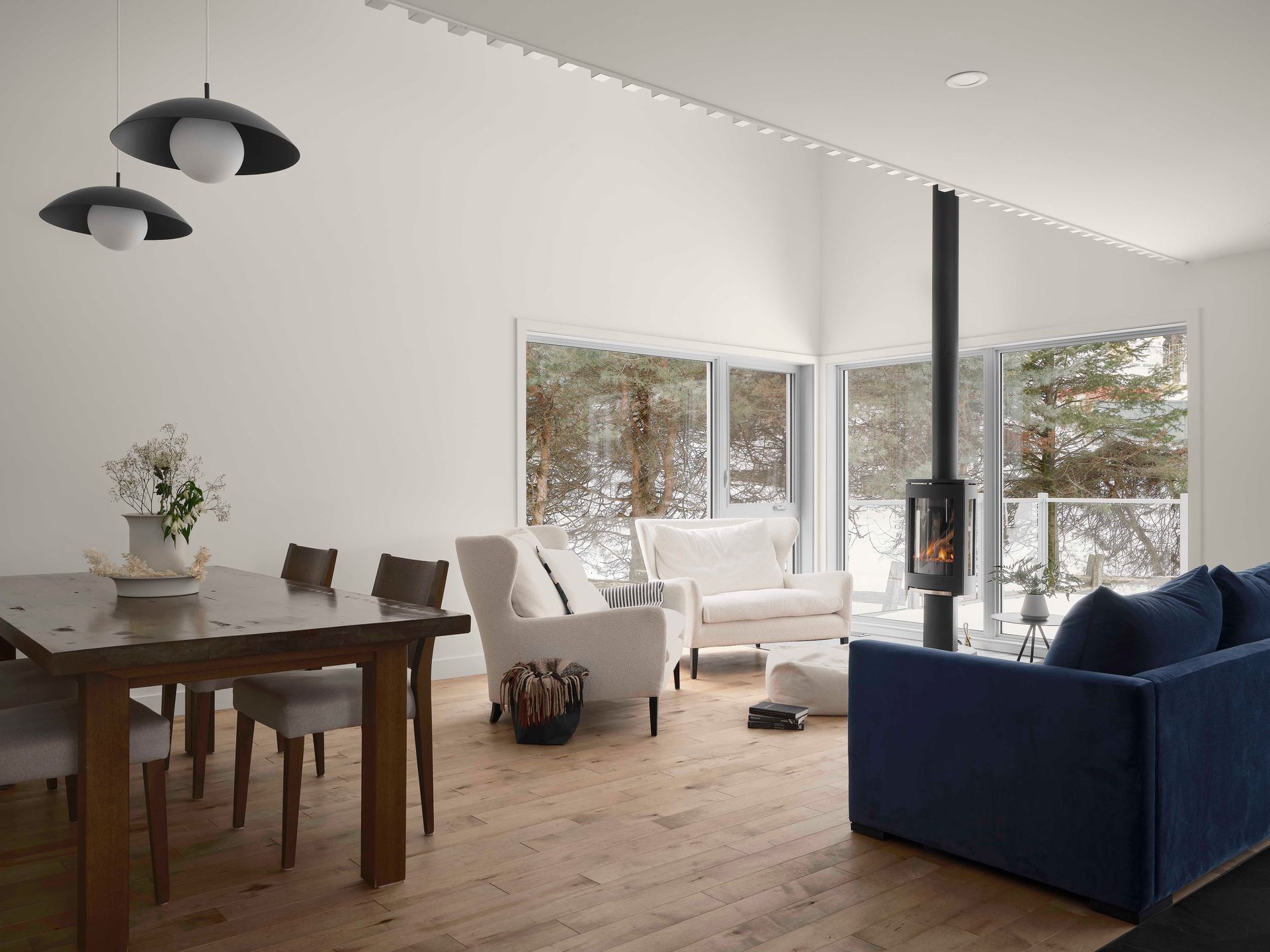Salon moderne avec coin repas, chaises blanches, canap&eacute; bleu et chemin&eacute;e noire donnant sur un paysage enneig&eacute;.
