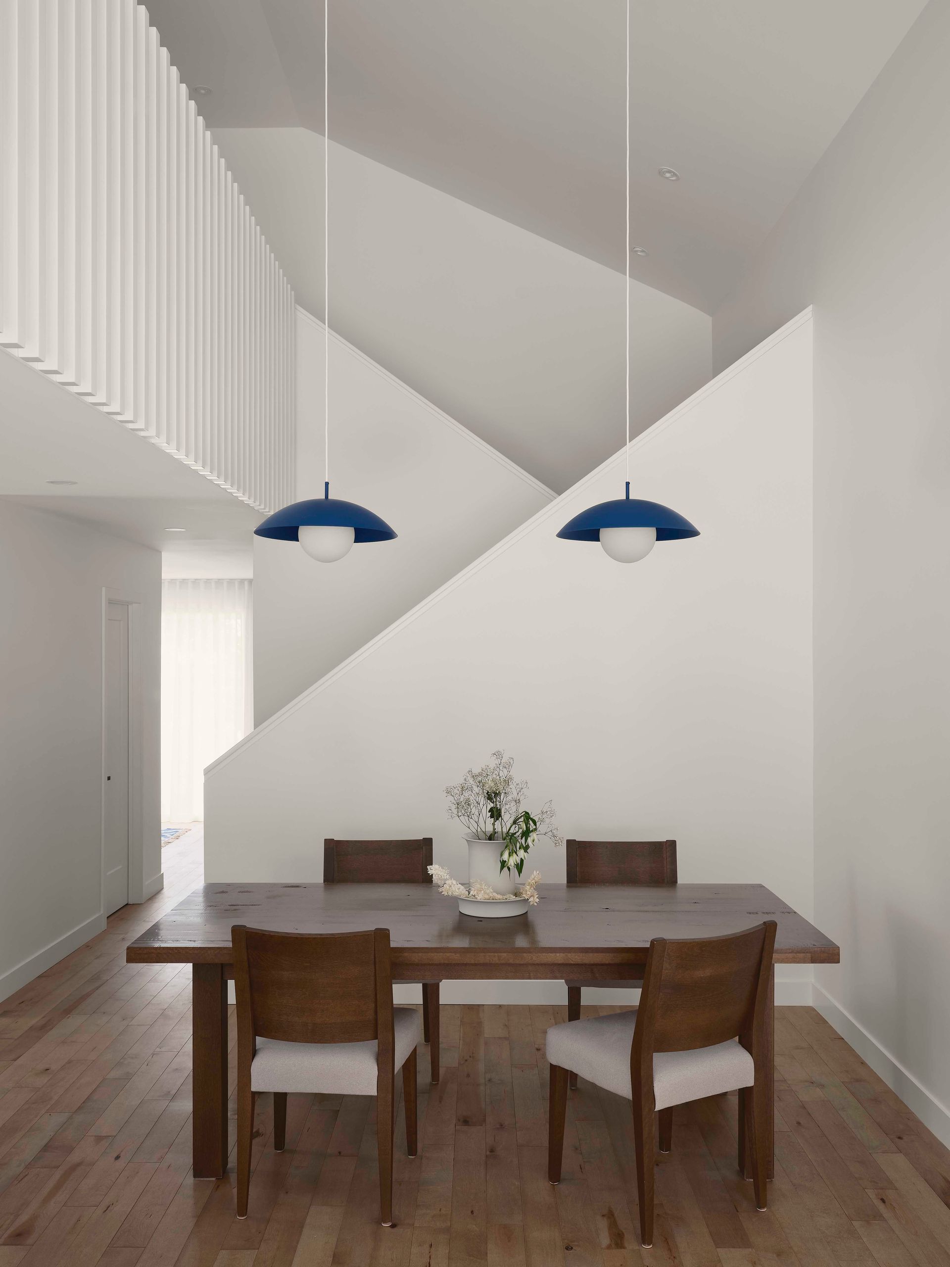 Une salle &agrave; manger minimaliste avec une table en bois fonc&eacute;, quatre chaises et deux suspensions bleues devant un mur d'escalier blanc.