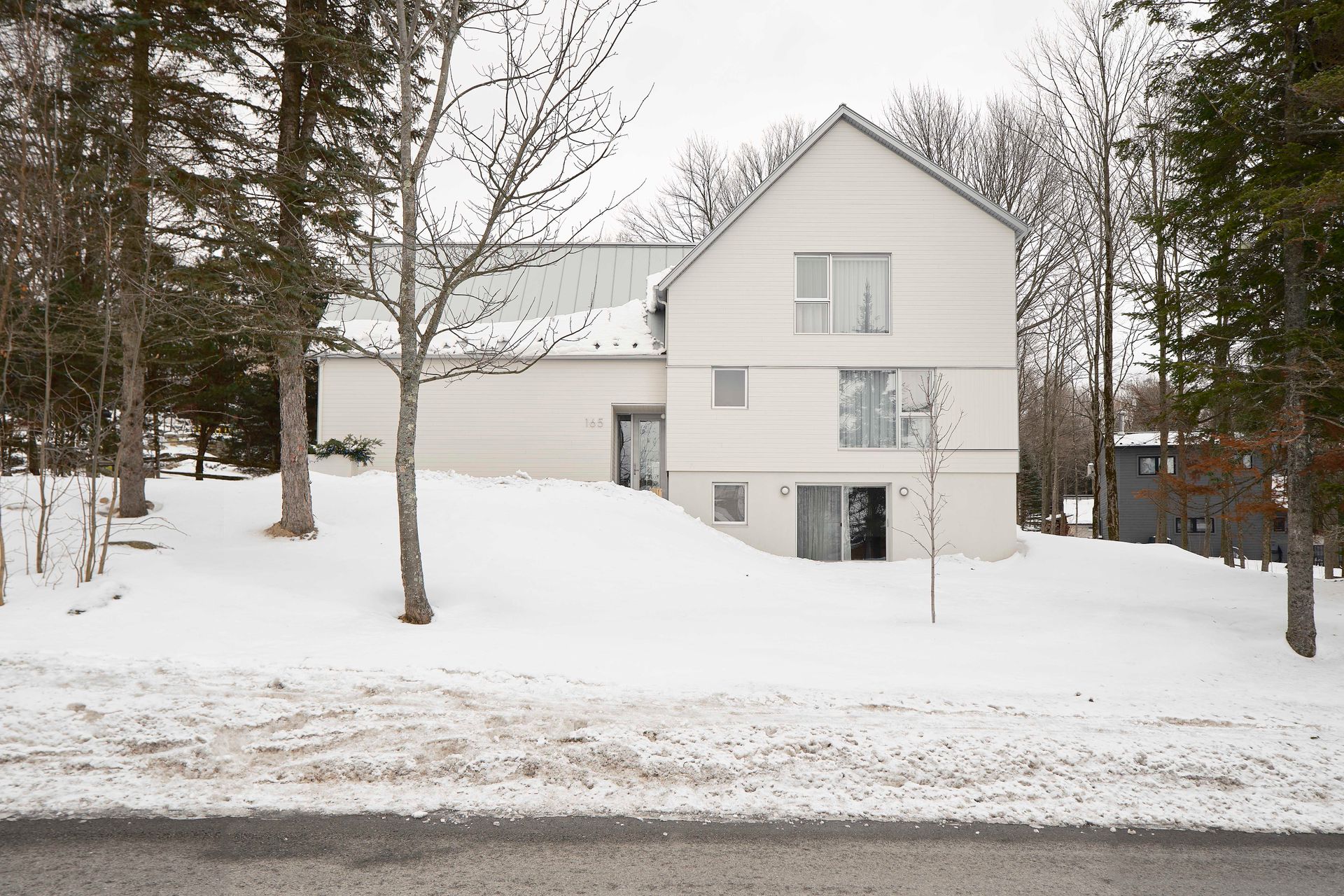 Une maison blanche moderne à deux étages, entourée de neige et d'arbres.