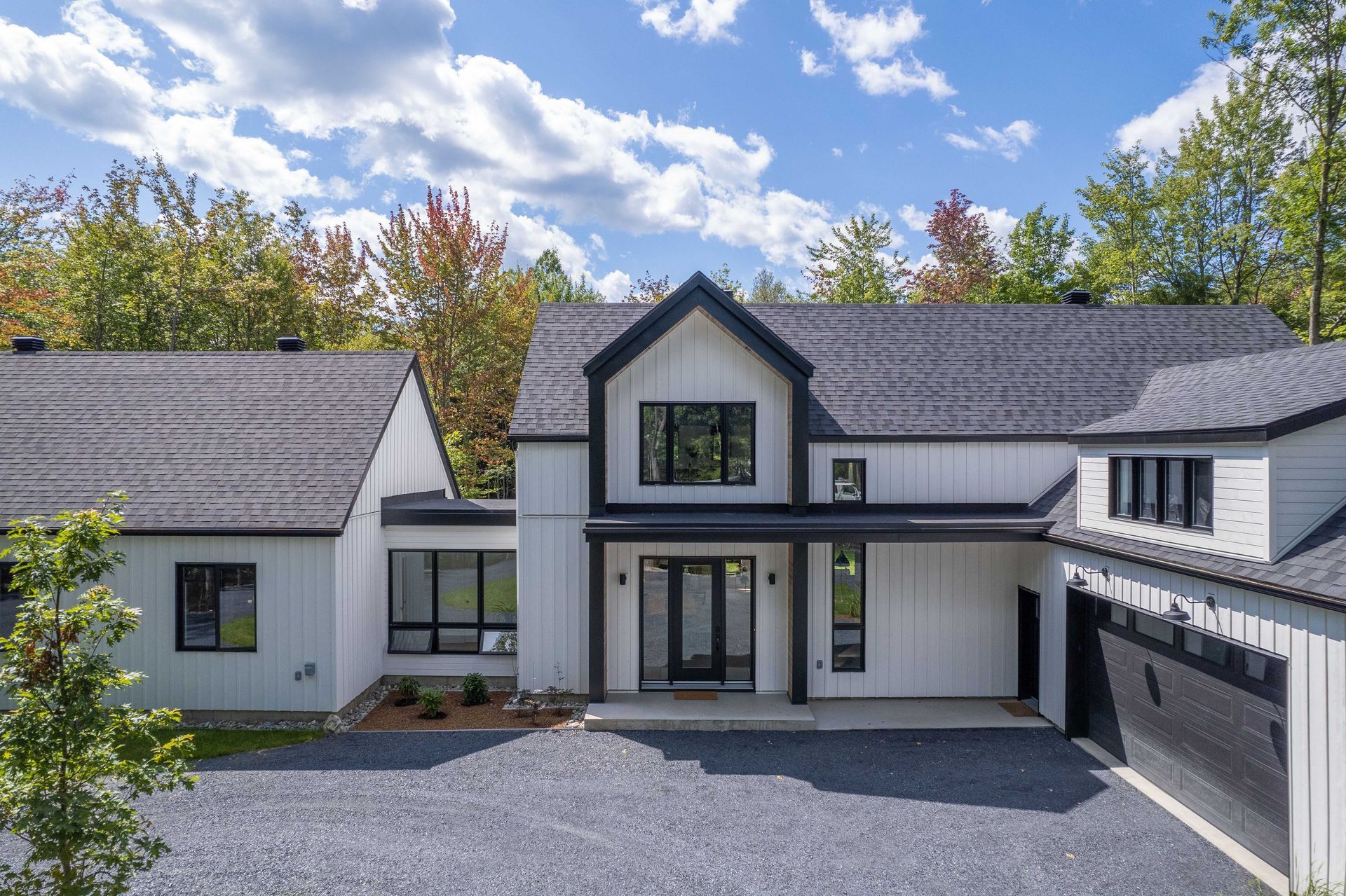 Une maison blanche moderne avec des boiseries noires et un toit gris foncé, agrémentée d'une allée de gravier et entourée d'arbres aux couleurs automnales.