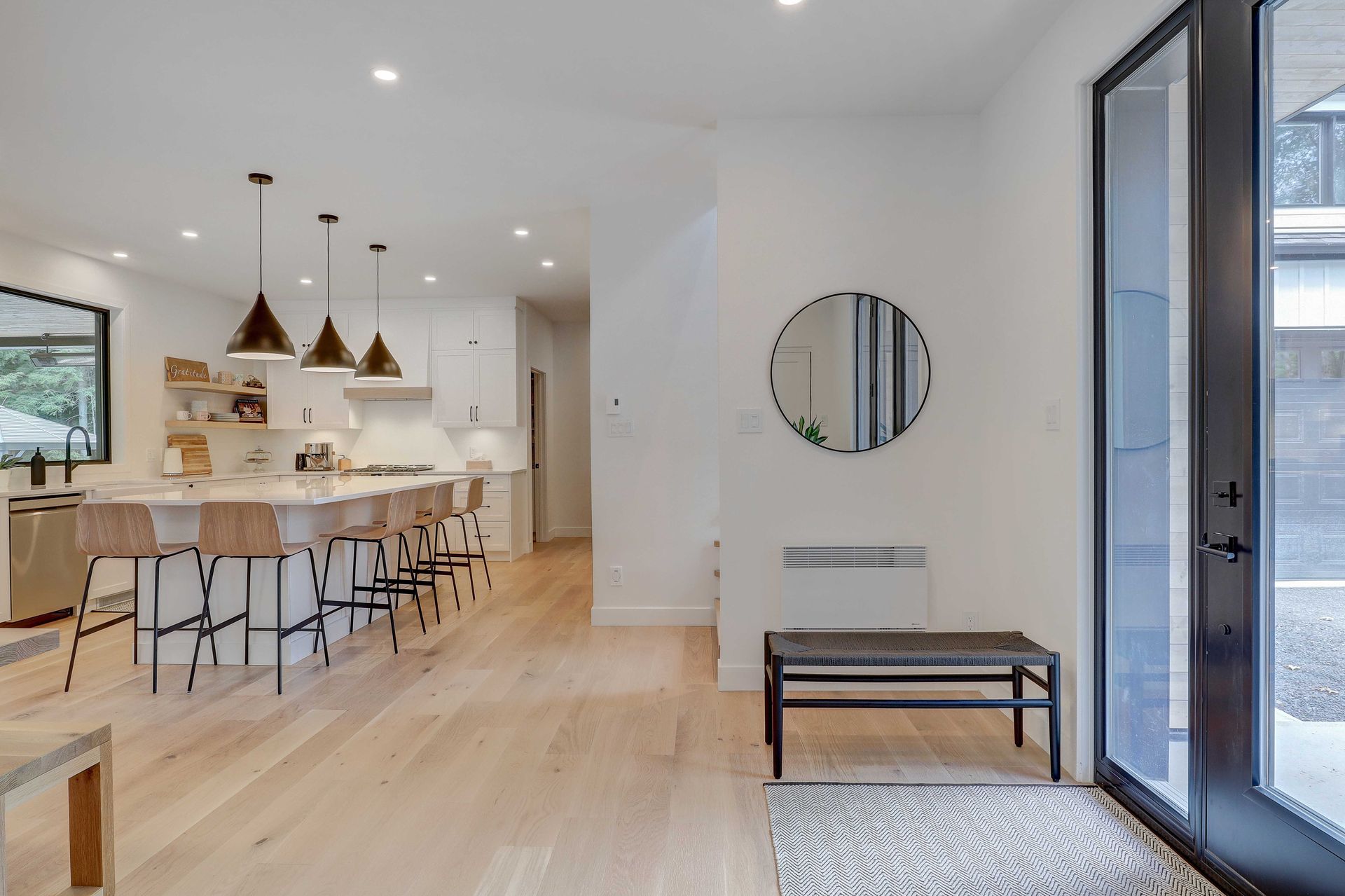 Cuisine moderne et lumineuse &agrave; aire ouverte avec un comptoir-bar, un plancher en bois et une entr&eacute;e ensoleill&eacute;e avec un banc et un miroir.