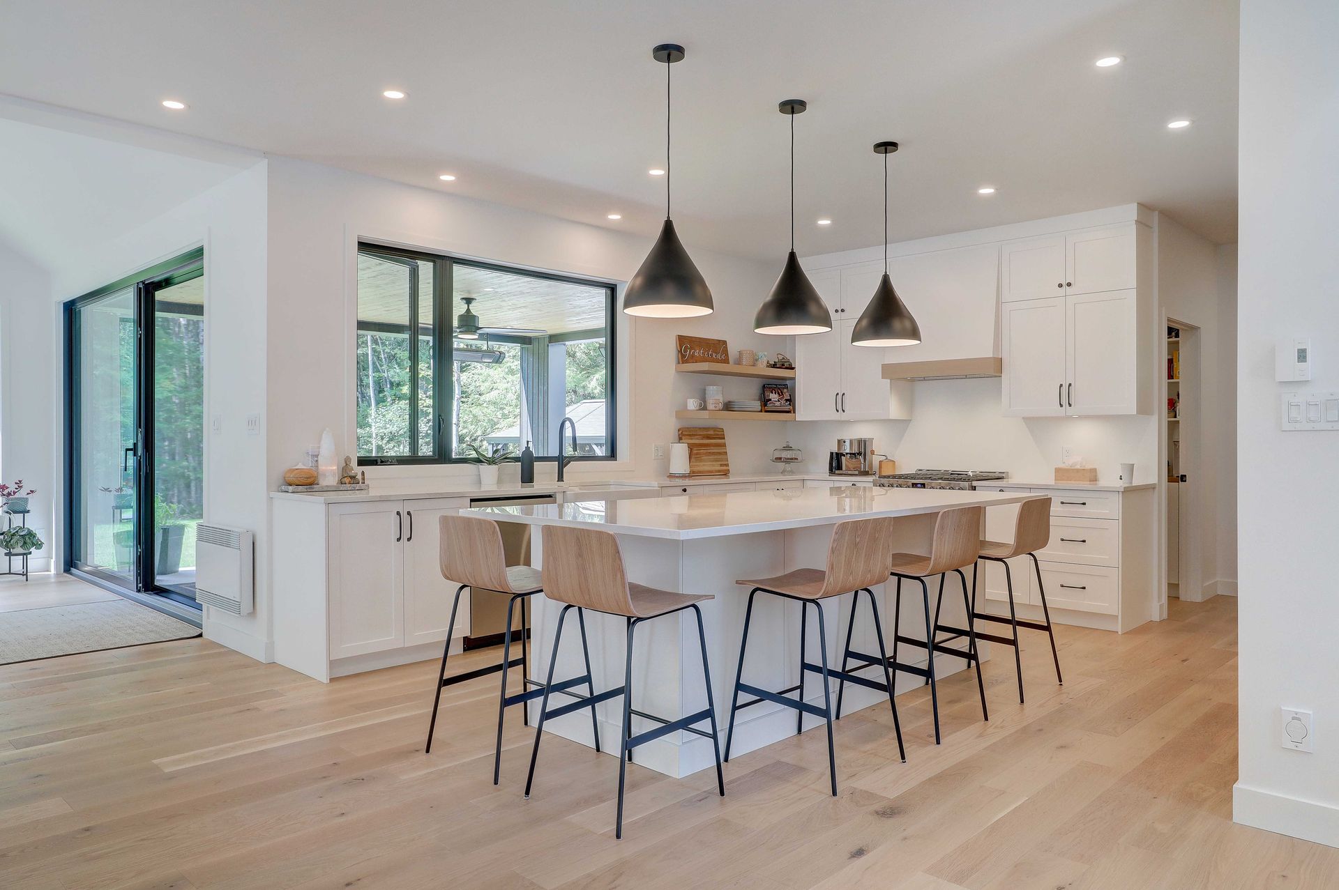 Une cuisine moderne et lumineuse avec des armoires blanches, un grand &icirc;lot avec des tabourets de bar en bois et trois suspensions lumineuses noires.