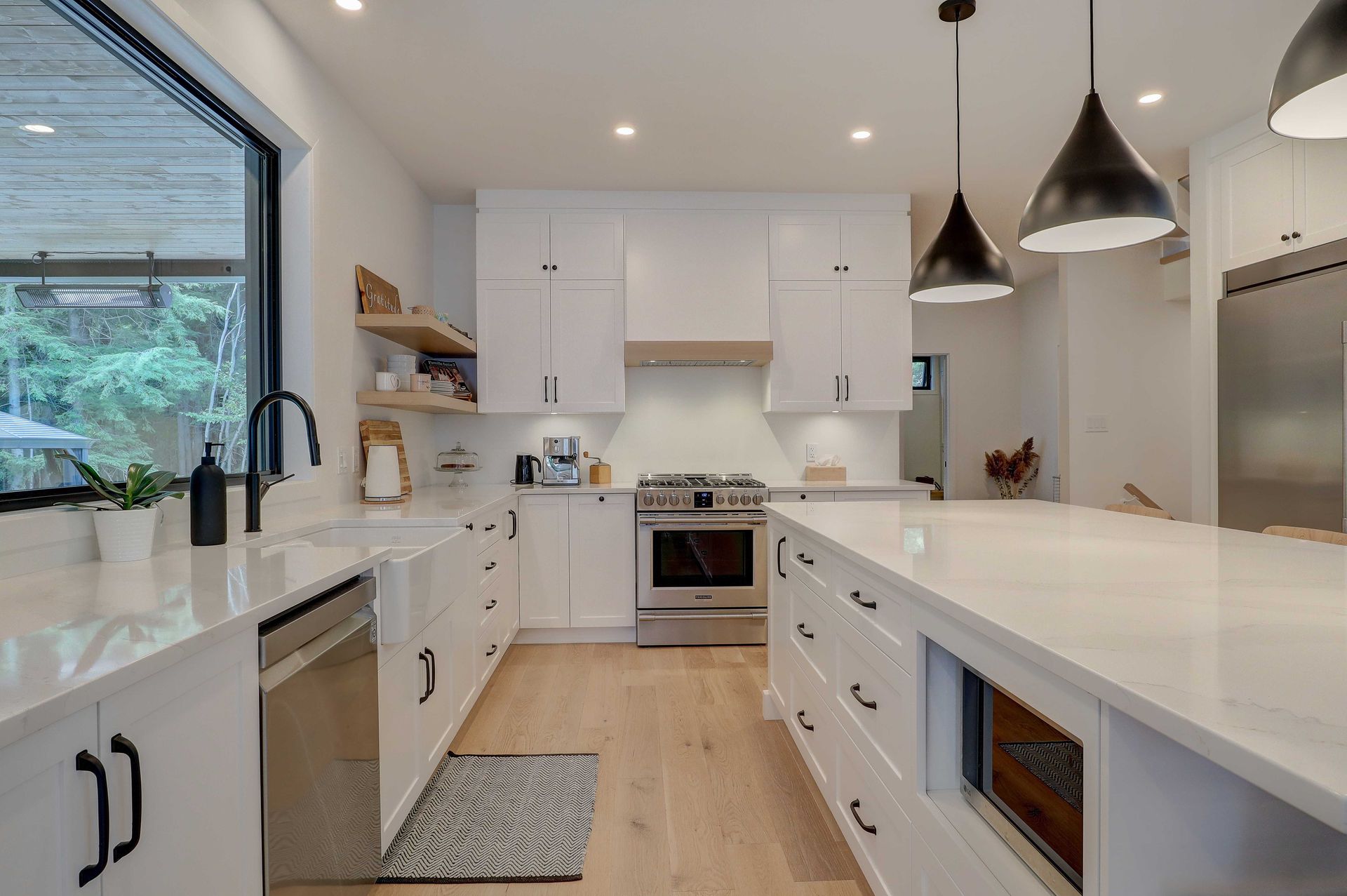 Cuisine blanche moderne avec appareils &eacute;lectrom&eacute;nagers en acier inoxydable, un grand &icirc;lot central, des suspensions lumineuses et une vue sur la verdure.