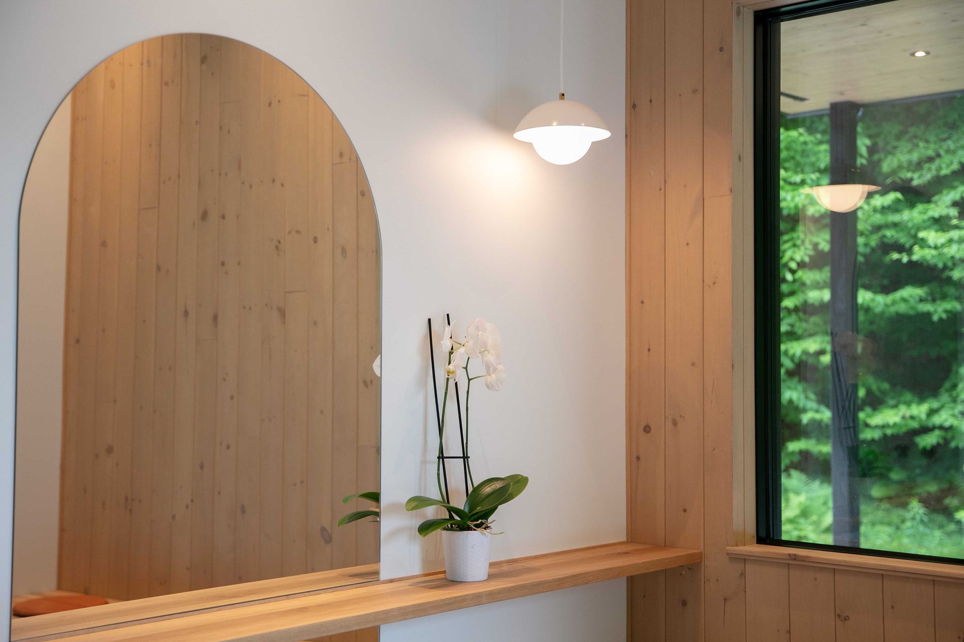 Un miroir arqu&eacute; refl&egrave;te des murs lambriss&eacute;s au-dessus d'une &eacute;tag&egrave;re en bois o&ugrave; repose une orchid&eacute;e en pot, &agrave; c&ocirc;t&eacute; d'une fen&ecirc;tre donnant sur les arbres.