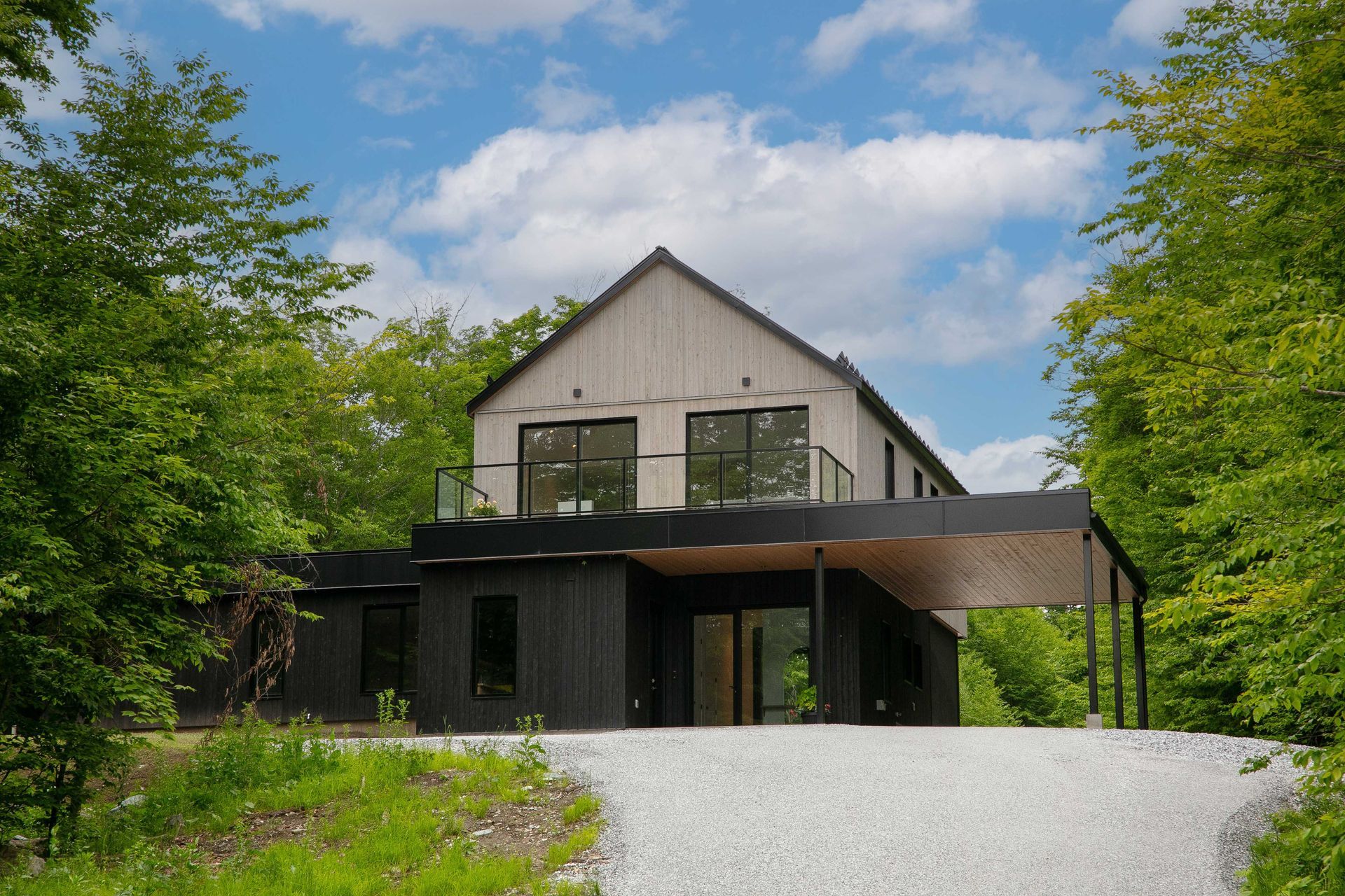 Une maison moderne à deux étages avec un bardage en bois clair et un rez-de-chaussée noir, nichée au milieu des arbres et dotée d'une allée de gravier.