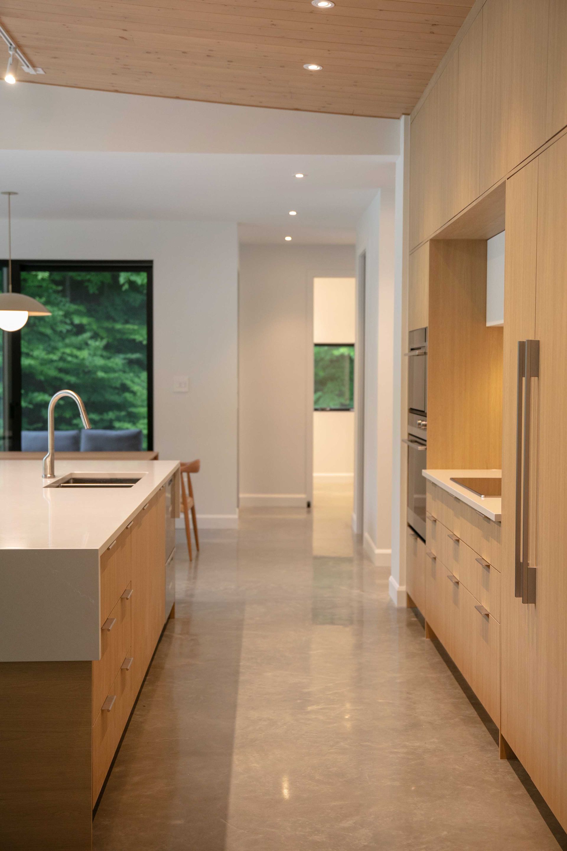 Un couloir-cuisine moderne avec des armoires en bois clair, des comptoirs blancs, des sols en b&eacute;ton et une vue sur une cour arri&egrave;re verdoyante.
