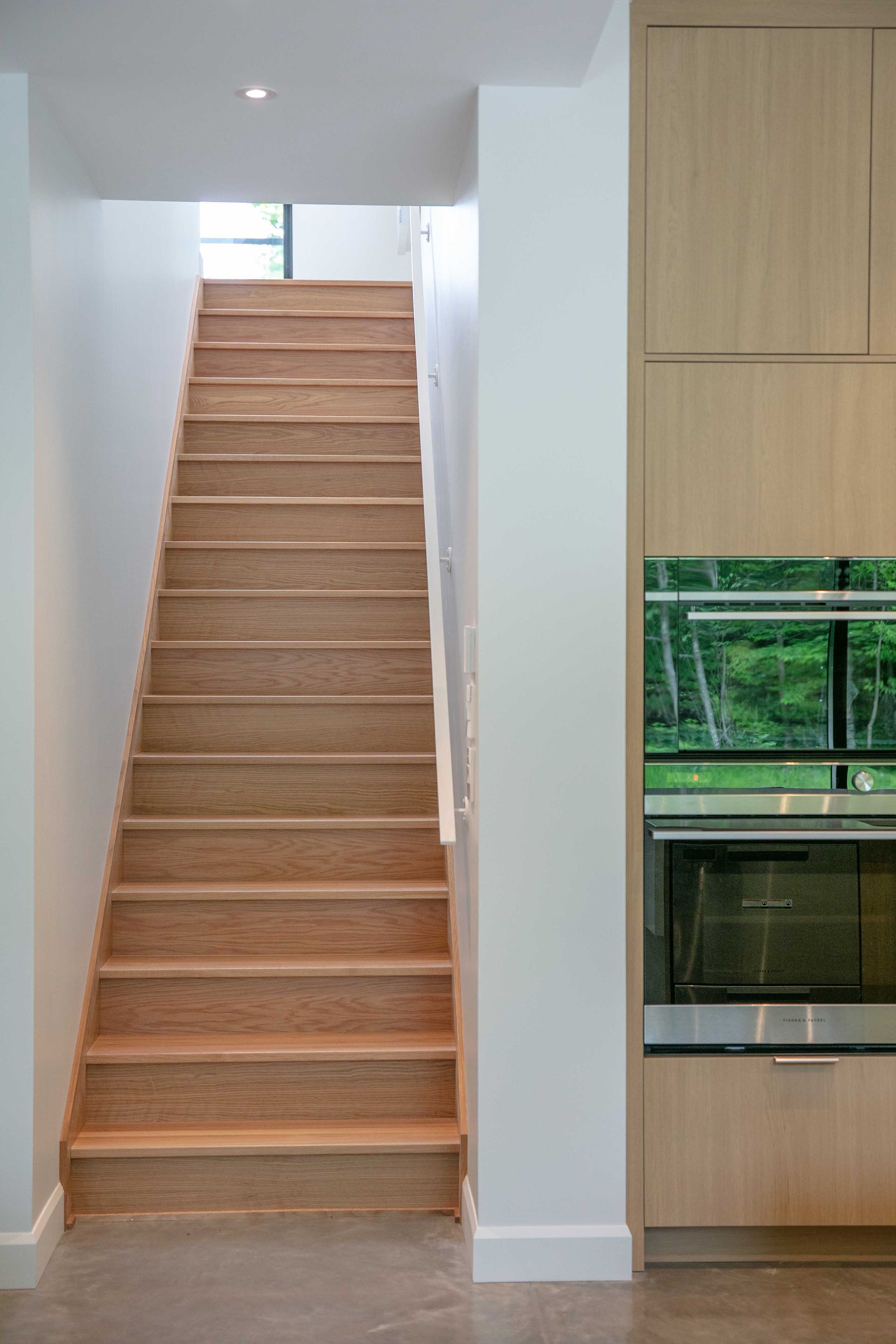 Un escalier droit en bois s'&eacute;l&egrave;ve entre des murs blancs, &agrave; c&ocirc;t&eacute; de placards en bois clair avec un four int&eacute;gr&eacute;.