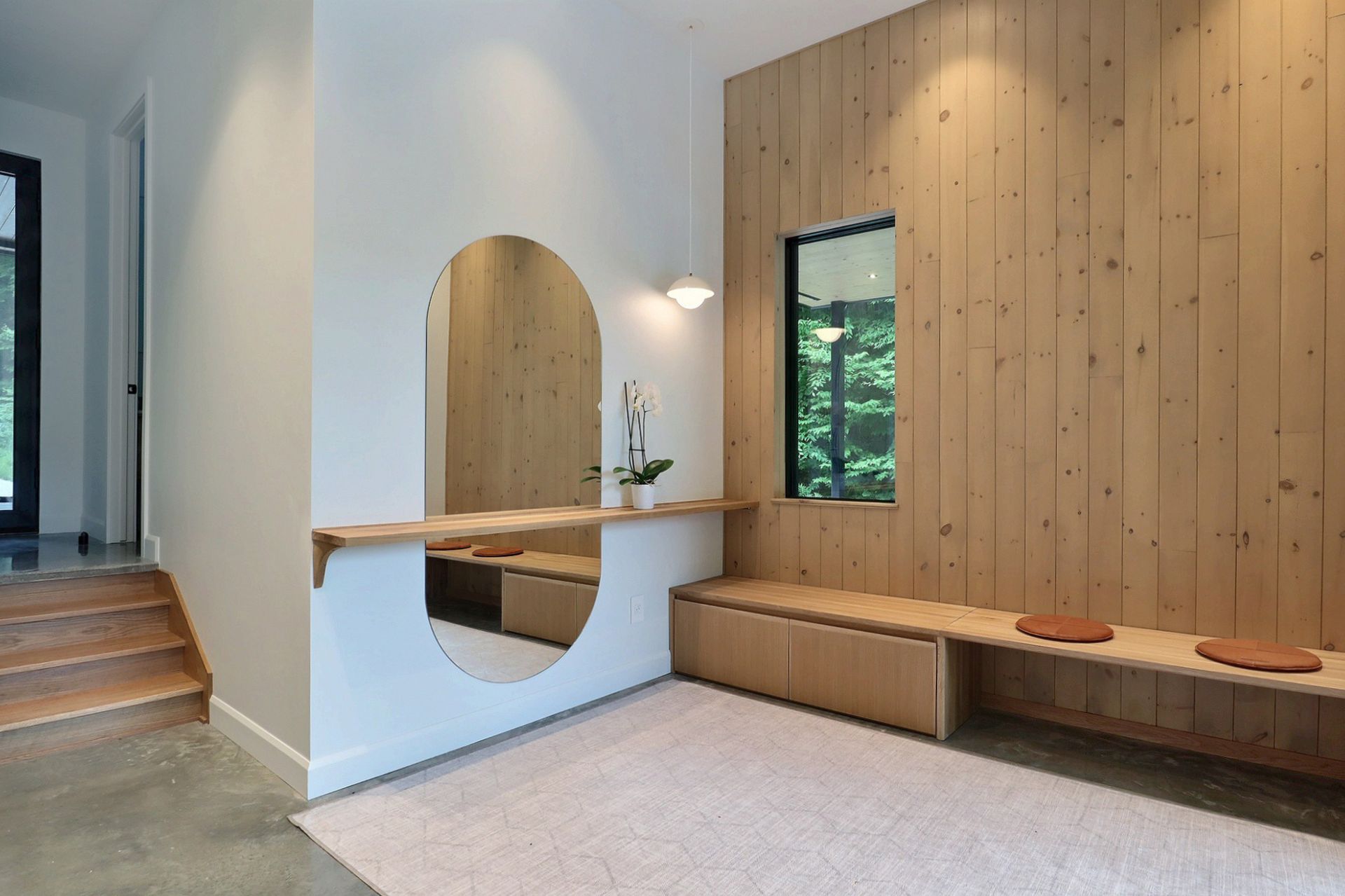 Une entr&eacute;e avec un mur en bois clair int&eacute;grant une banquette, un miroir rond avec une &eacute;tag&egrave;re, un tapis et une suspension lumineuse.