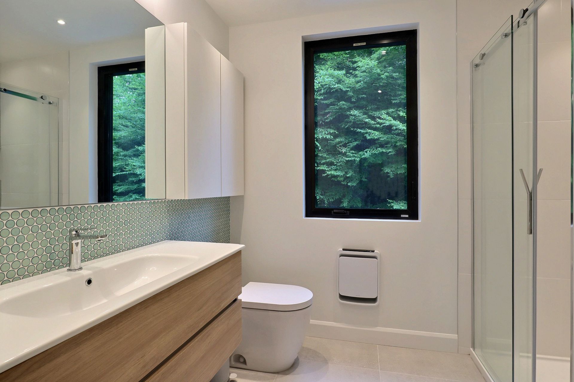 Une salle de bains moderne et lumineuse avec un meuble-lavabo en bois clair, une cr&eacute;dence en mosa&iuml;que, des toilettes blanches et une cabine de douche en verre.