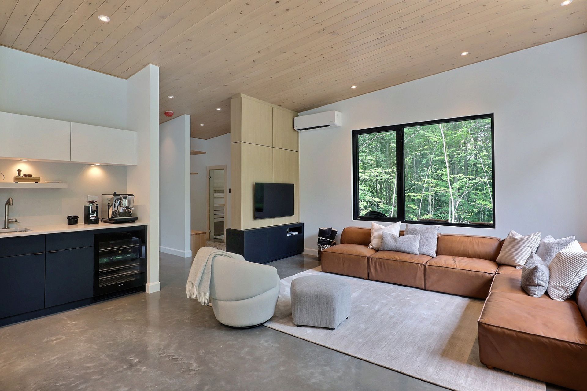 Un salon moderne avec un canap&eacute; d'angle en cuir marron, un fauteuil pivotant blanc, une kitchenette et une grande fen&ecirc;tre donnant sur les arbres.
