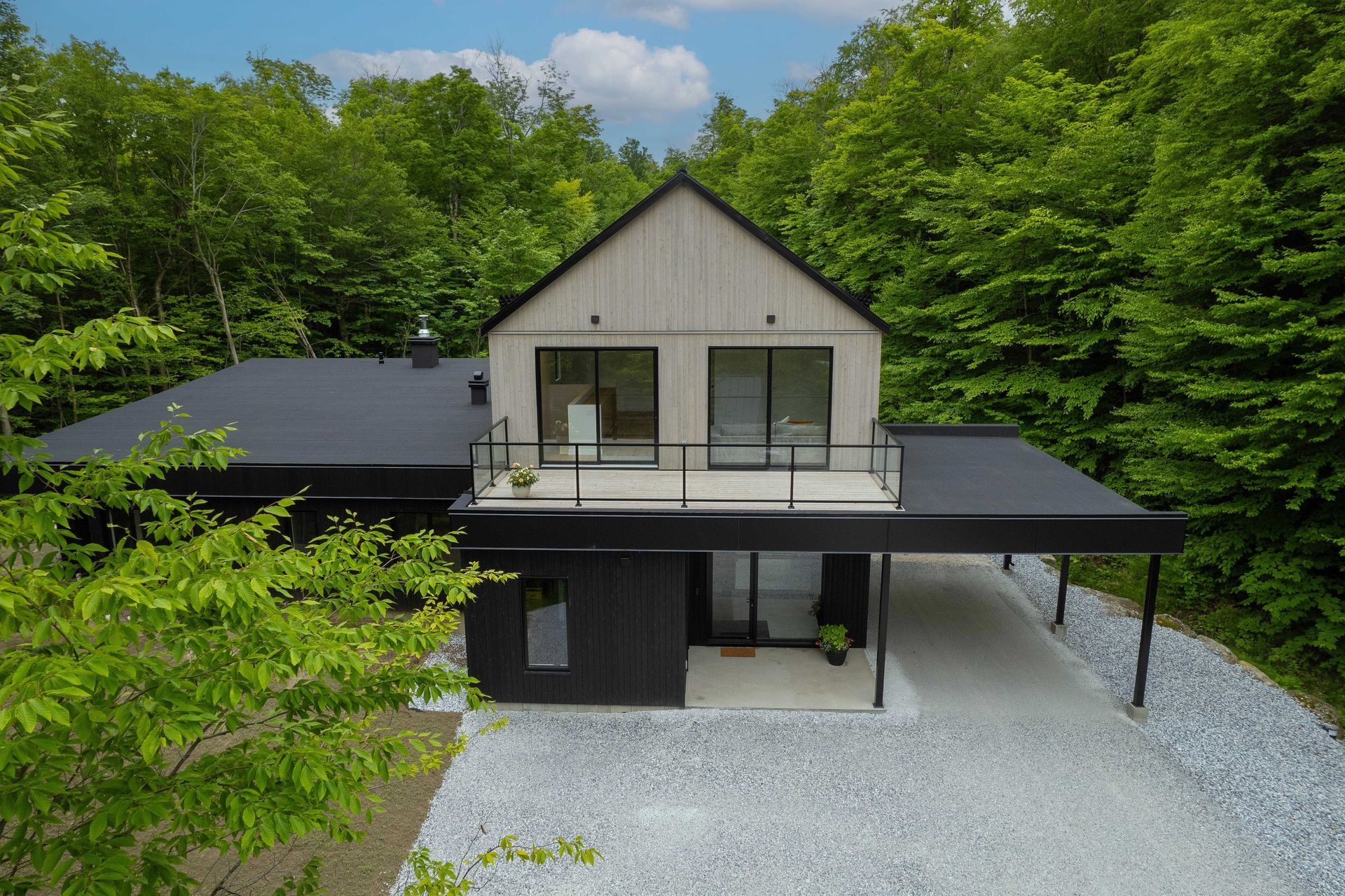 Une maison moderne à deux étages avec un bardage en bois clair et un rez-de-chaussée noir, nichée au milieu des arbres et dotée d'une allée de gravier.