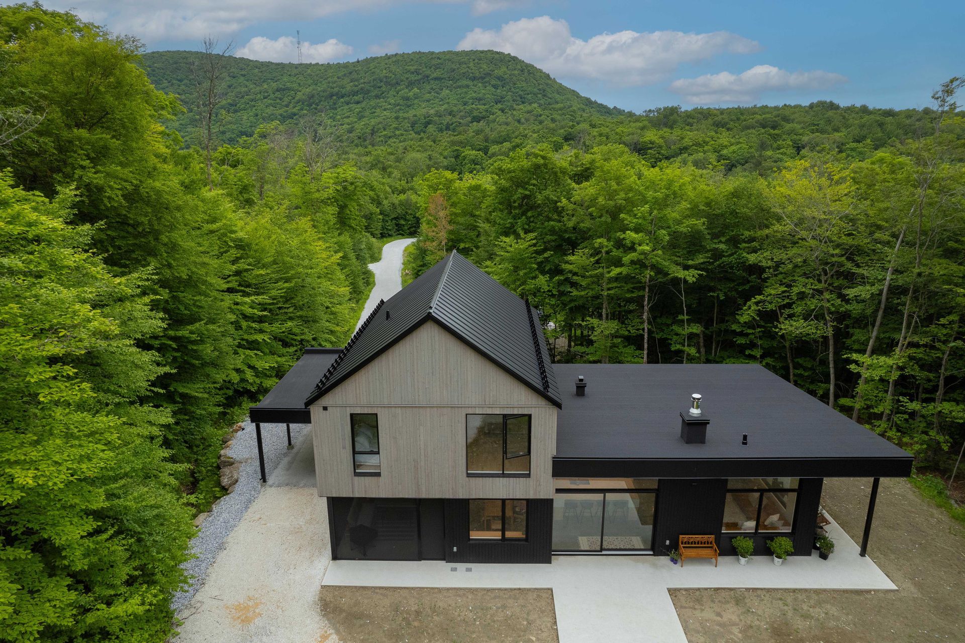 Vue a&eacute;rienne d'une maison moderne &agrave; deux &eacute;tages, avec un toit noir et un bardage en bois clair, nich&eacute;e dans une for&ecirc;t luxuriante et verdoyante.