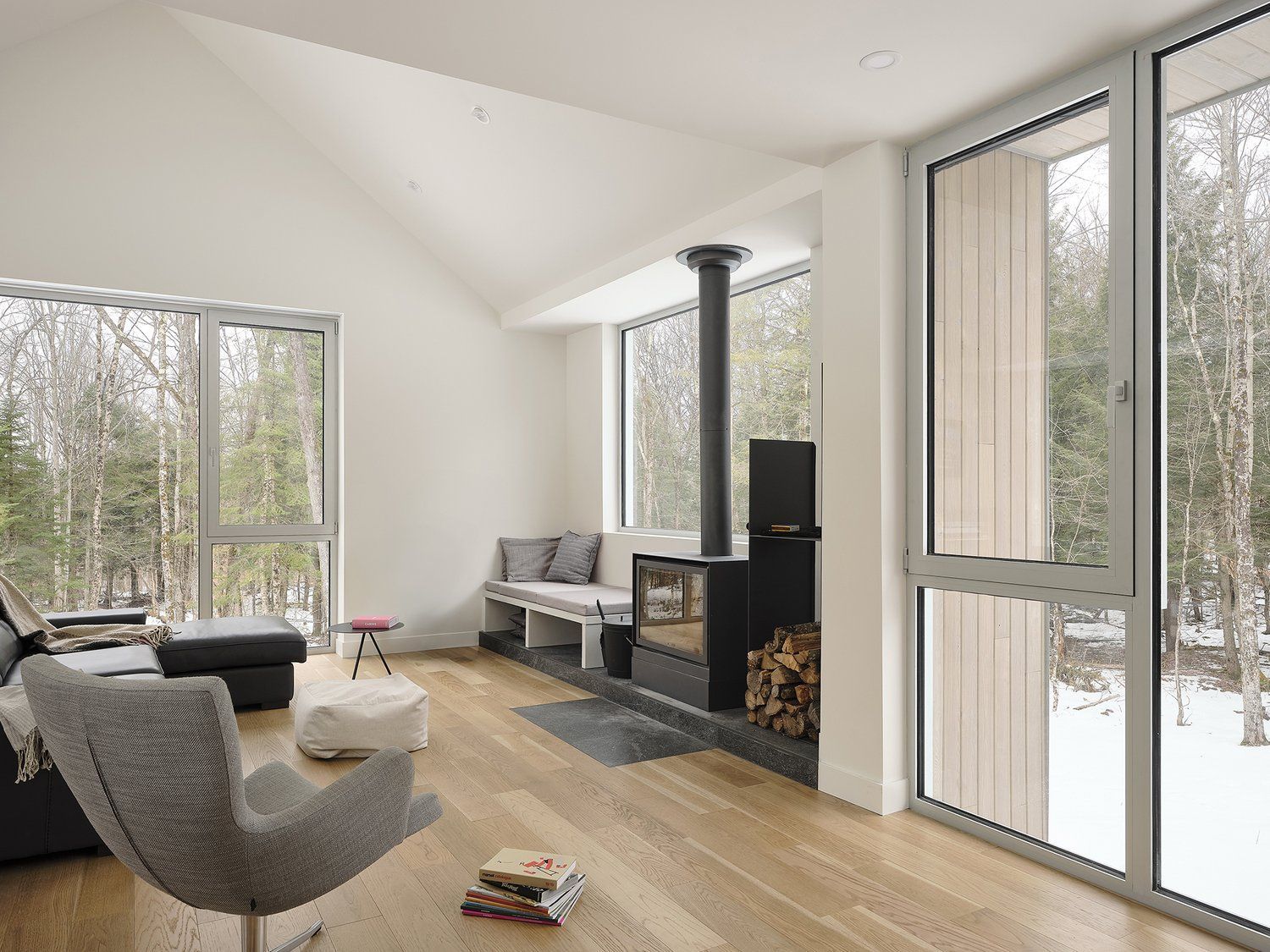 Salon moderne et lumineux avec parquet, po&ecirc;le &agrave; bois et grandes fen&ecirc;tres donnant sur un paysage bois&eacute; enneig&eacute;.