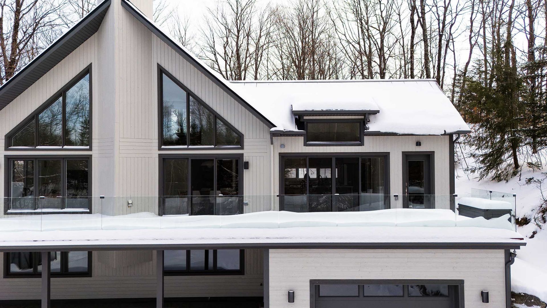 Modern house with a pitched roof and large windows, partially covered in snow against a backdrop of bare winter trees.