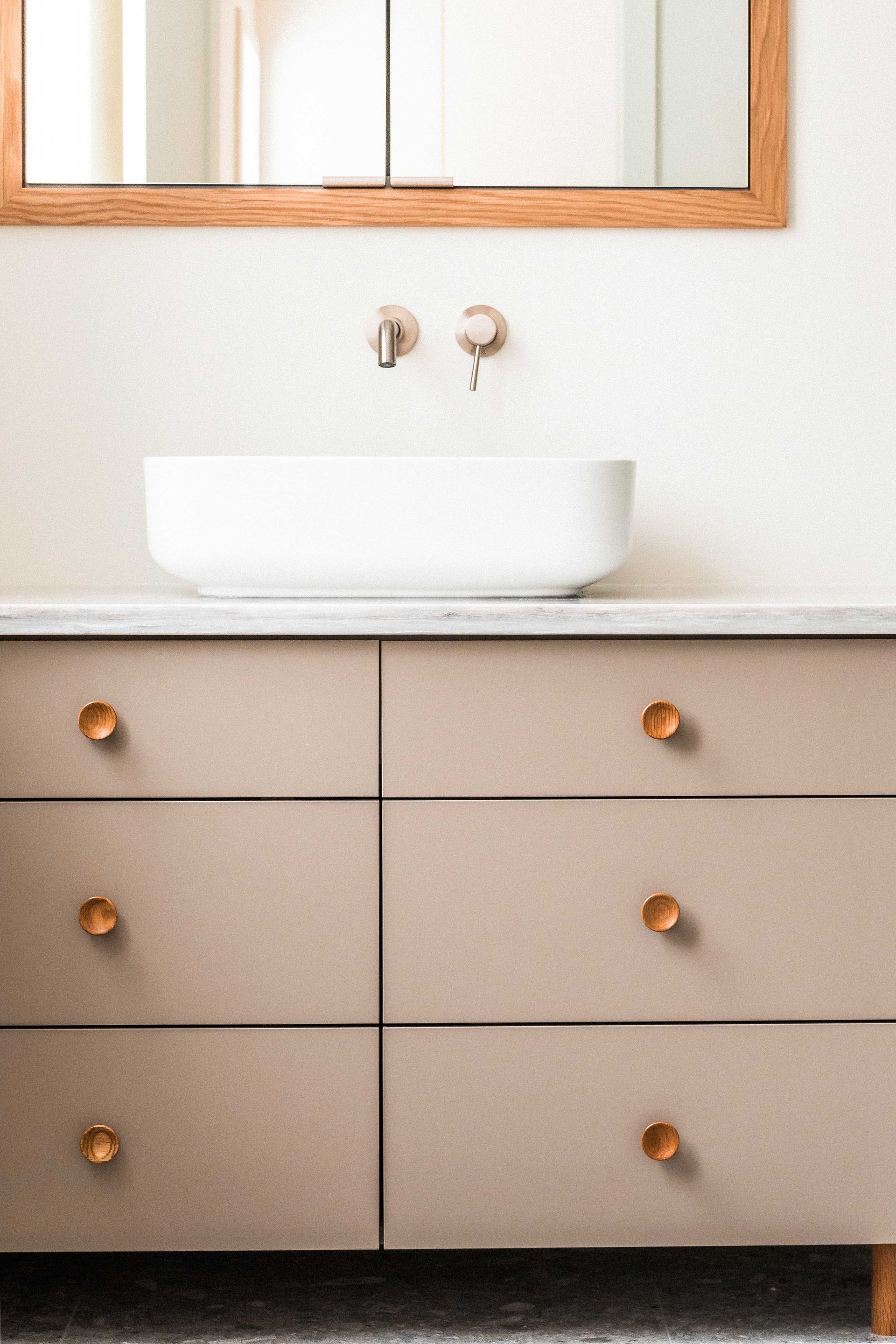 Un meuble-lavabo moderne avec six tiroirs beiges, des poign&eacute;es rondes en laiton, un lavabo vasque blanc et un miroir encadr&eacute; de bois.