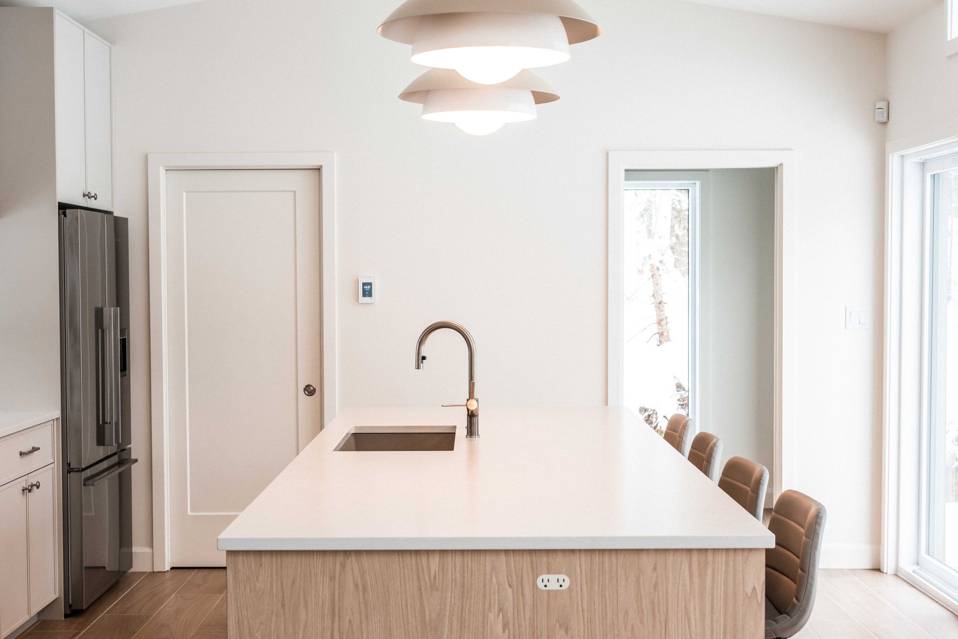 Un &icirc;lot de cuisine lumineux, avec une base en bois clair et un plan de travail blanc, tr&ocirc;ne au centre, devant deux suspensions lumineuses modernes.