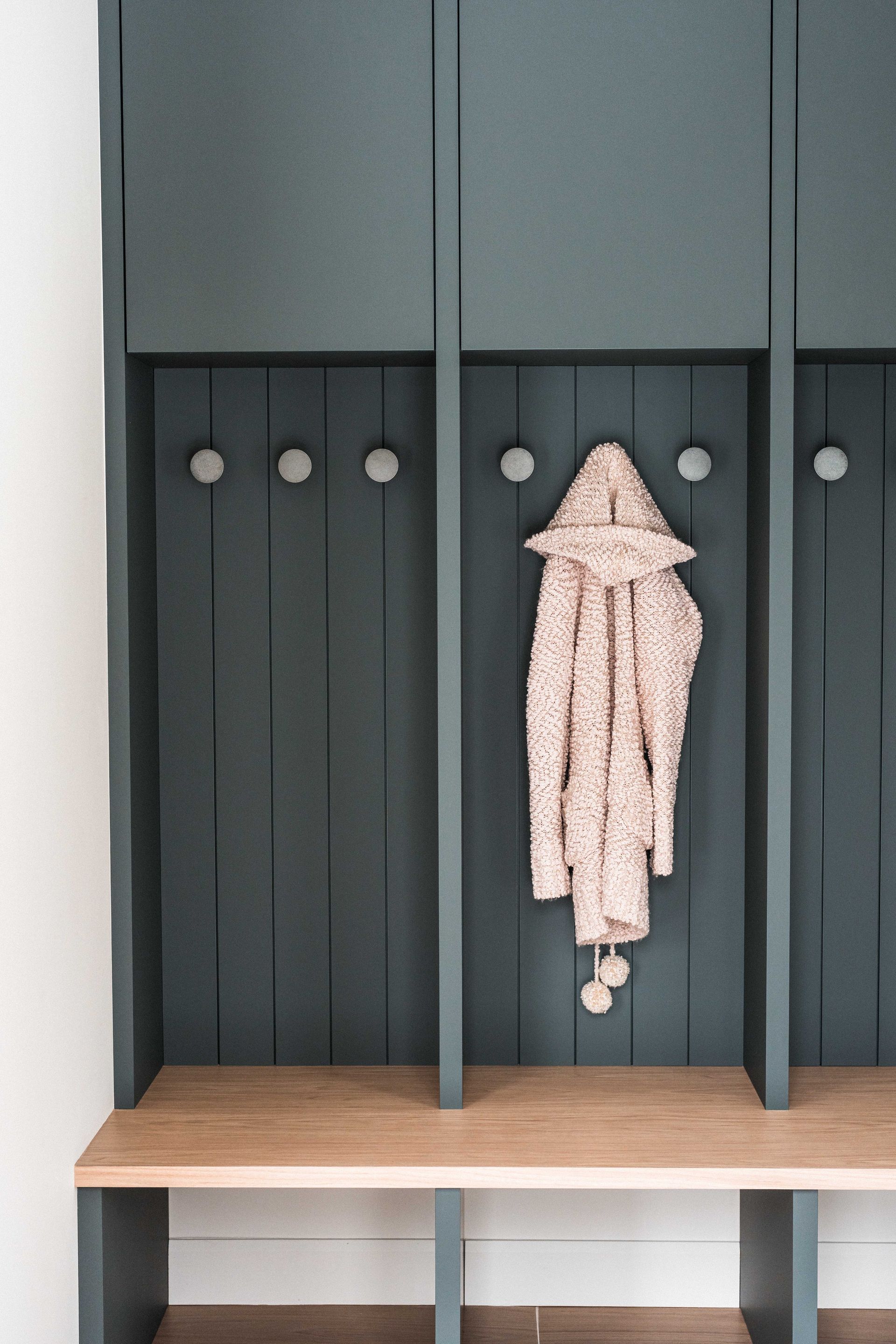 Casiers vert fonc&eacute; dans le vestibule, avec un banc en bois et des crochets; un crochet retient un cardigan &agrave; capuche textur&eacute; de couleur claire.