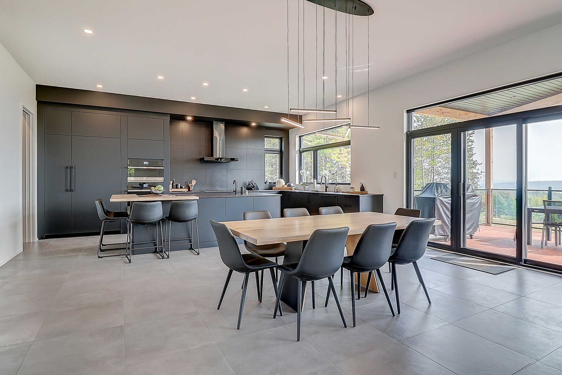 Une cuisine et une salle &agrave; manger modernes &agrave; aire ouverte avec des armoires noir mat, une table &agrave; manger en bois et de grandes portes coulissantes.