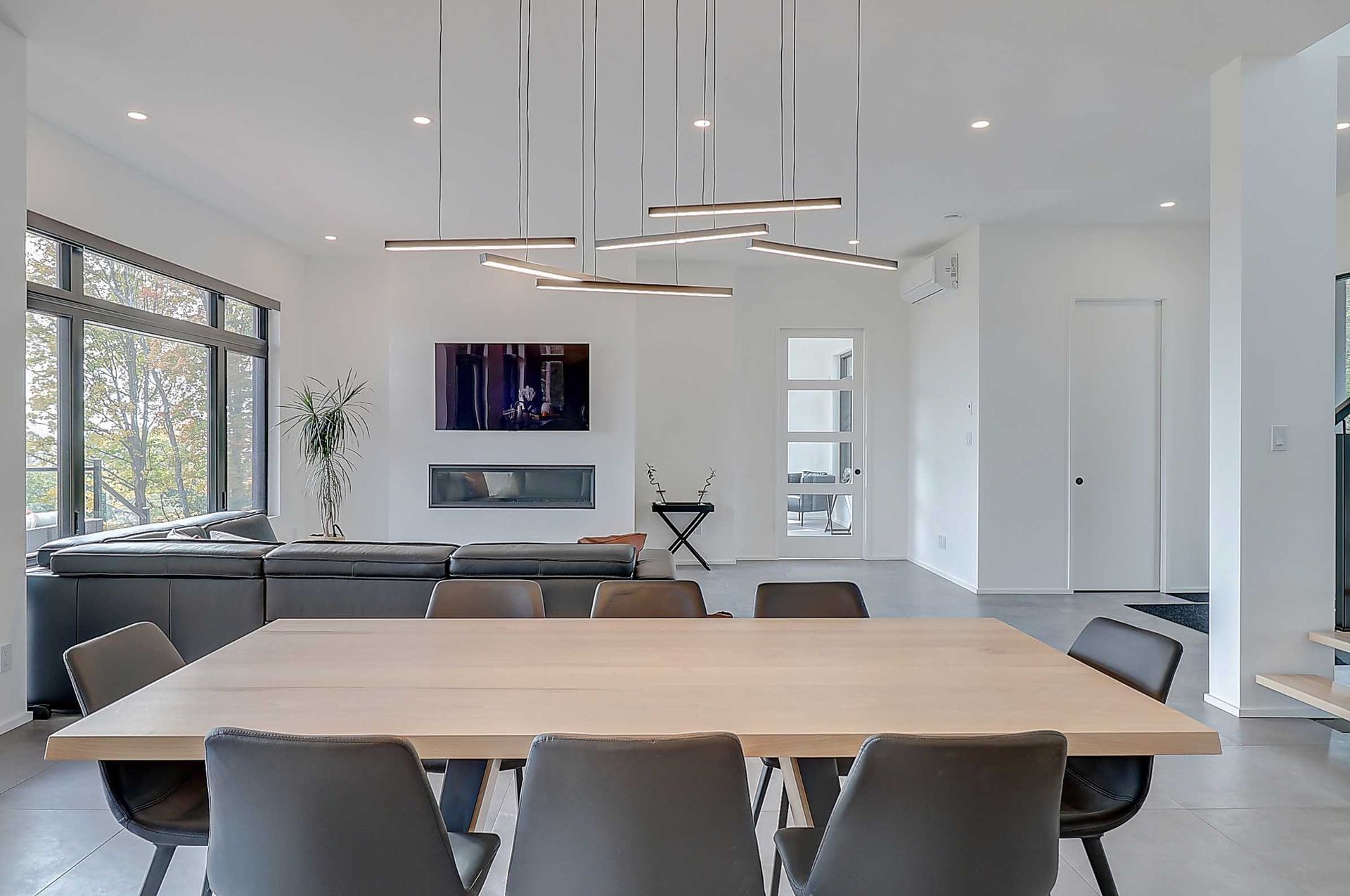 Un espace de vie et salle &agrave; manger lumineux et moderne, &agrave; aire ouverte, avec une table en bois clair, des chaises grises et un lustre g&eacute;om&eacute;trique.