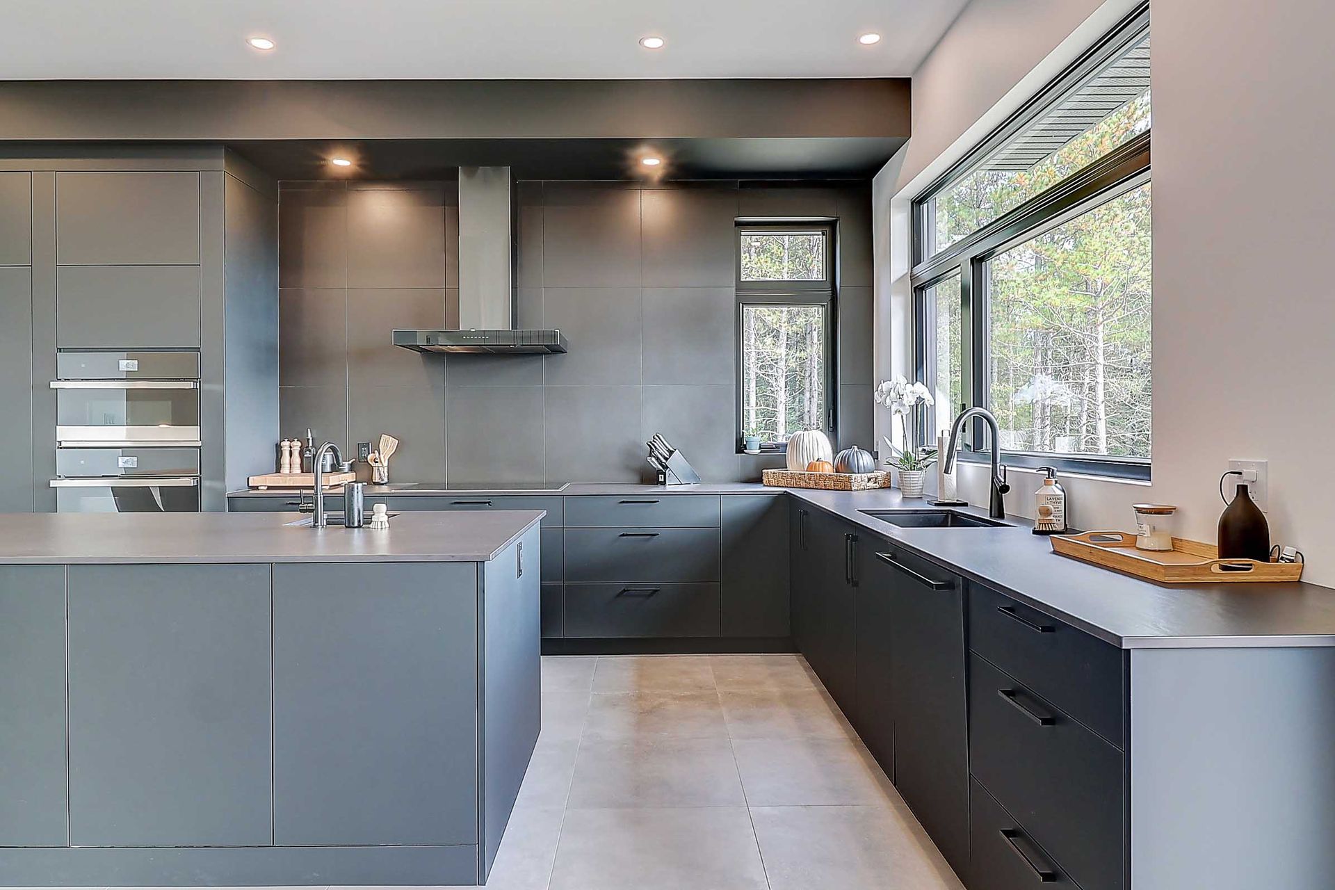 Une cuisine moderne et minimaliste avec des armoires &agrave; panneaux plats gris fonc&eacute;, un grand &icirc;lot central et une large fen&ecirc;tre.