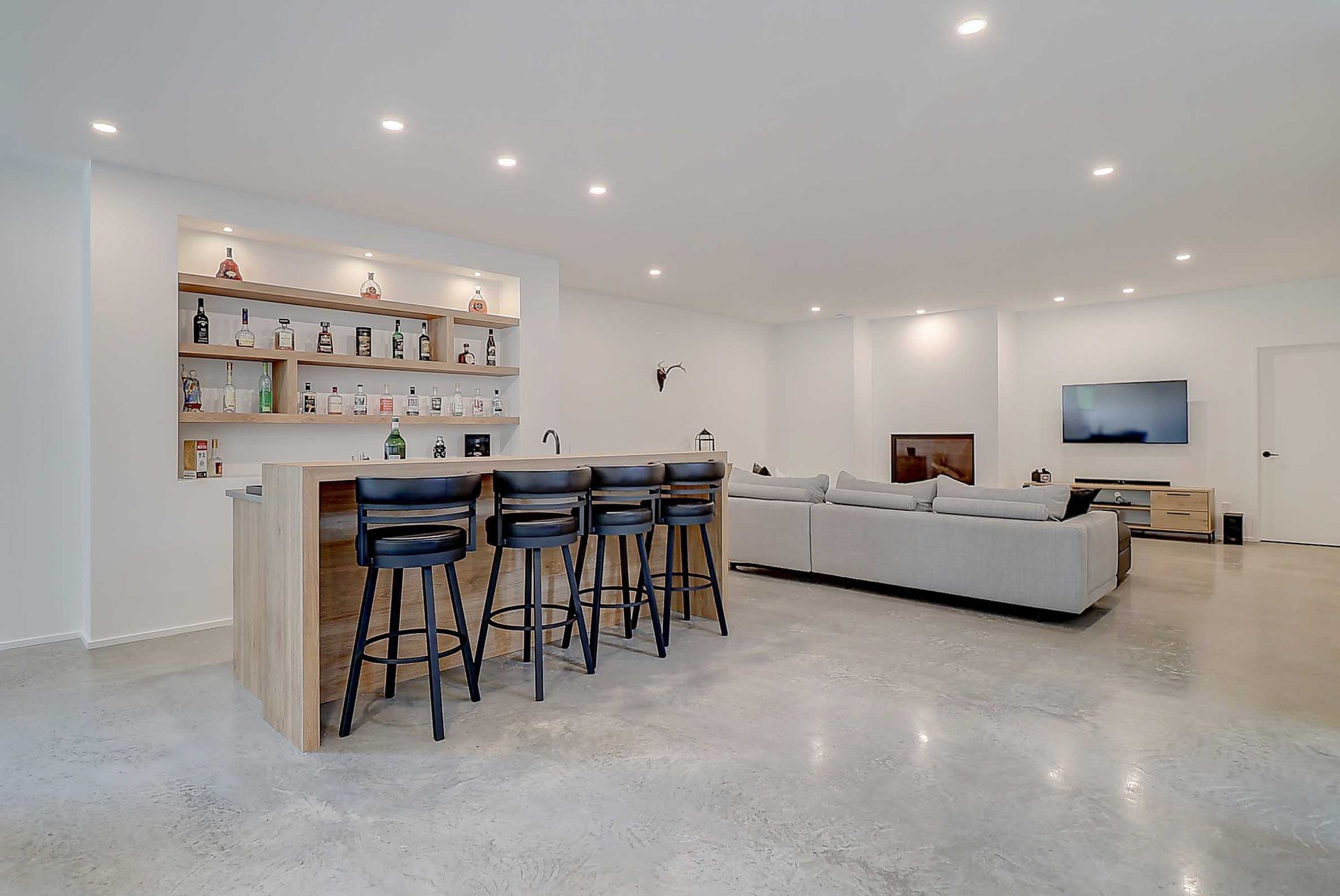 Sous-sol am&eacute;nag&eacute; moderne avec un bar en bois, quatre tabourets, des &eacute;tag&egrave;res encastr&eacute;es, un canap&eacute; gris et un sol en b&eacute;ton clair.