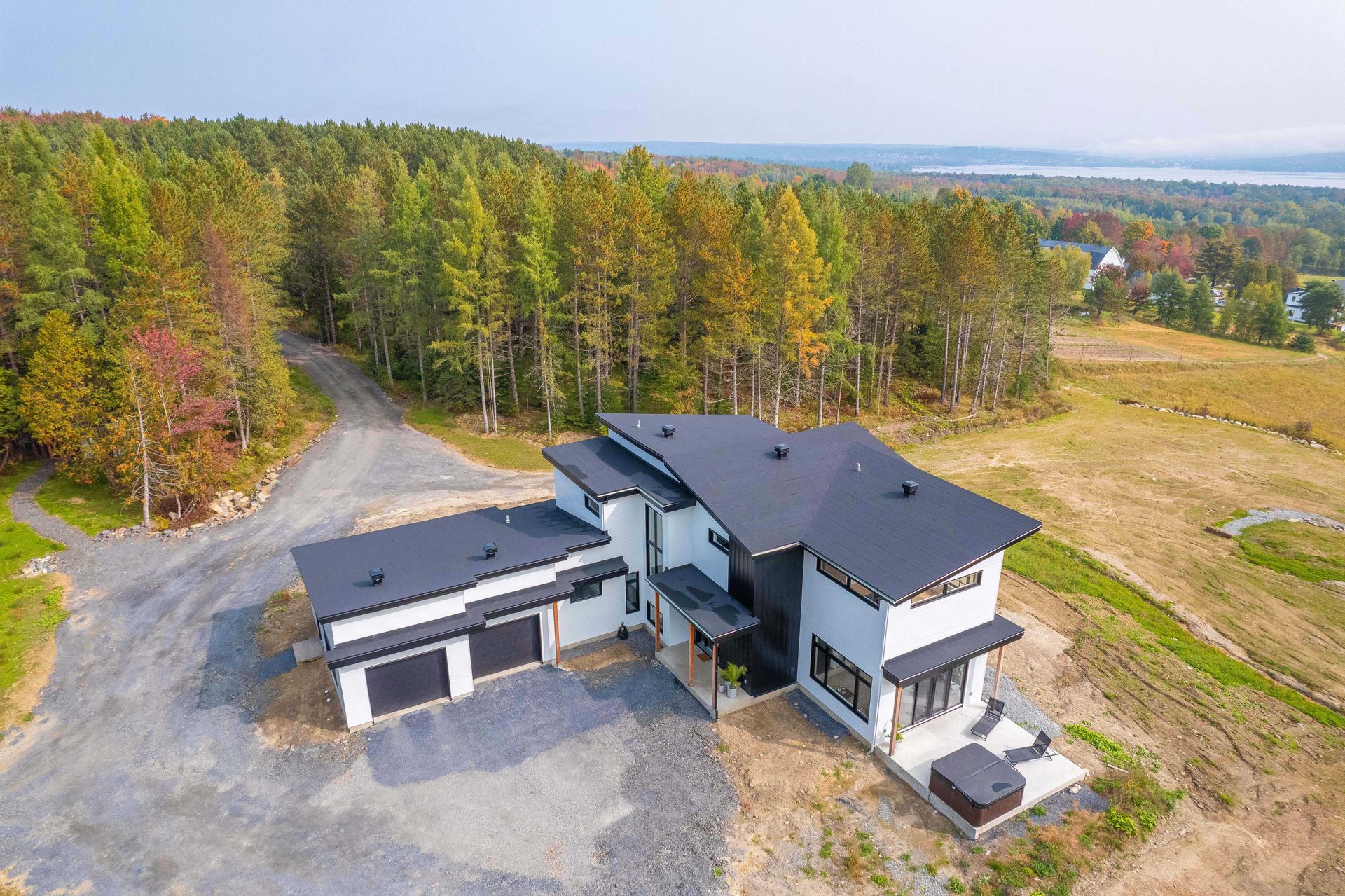 Vue a&eacute;rienne d'une maison moderne &agrave; deux &eacute;tages, avec un toit noir et des murs blancs, situ&eacute;e pr&egrave;s d'une for&ecirc;t et d'un champ.