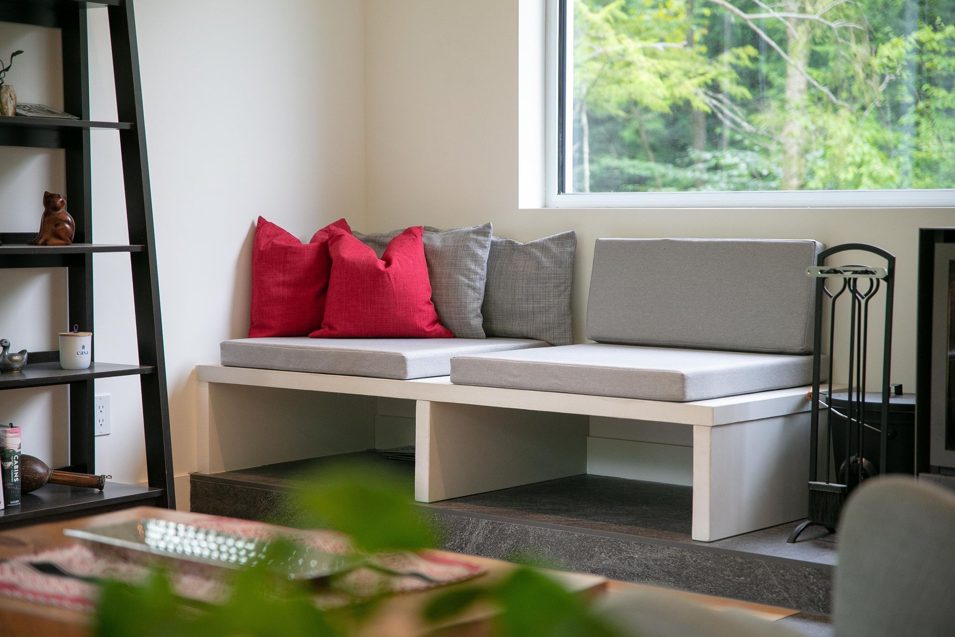 Un banc en bois blanc, avec des coussins d'assise gris et des coussins d&eacute;coratifs rouges, est plac&eacute; sous une fen&ecirc;tre, &agrave; c&ocirc;t&eacute; d'une &eacute;tag&egrave;re noire.