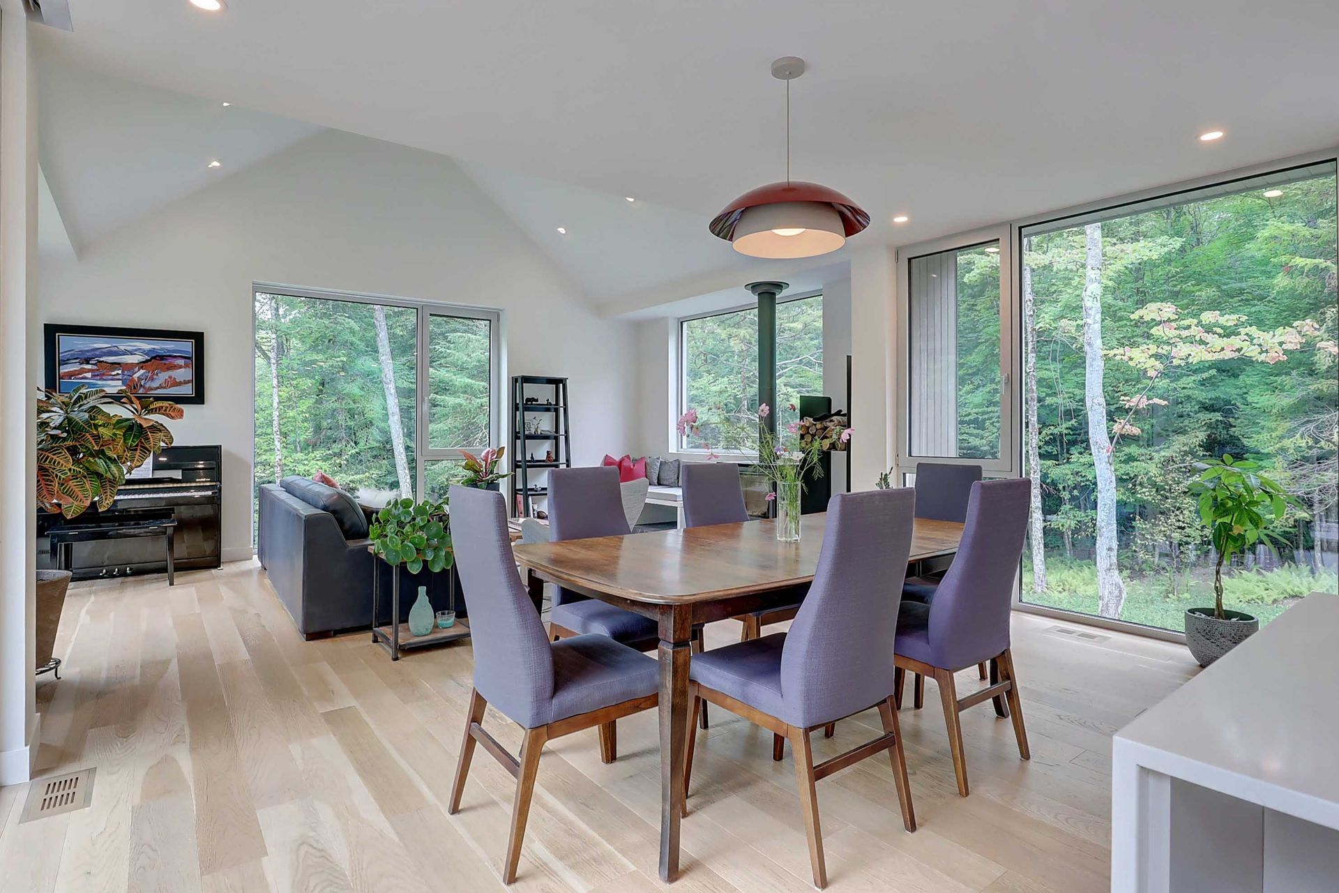 Salle &agrave; manger moderne &agrave; aire ouverte avec une table en bois, des chaises violettes, un parquet clair et de grandes fen&ecirc;tres donnant sur les bois.