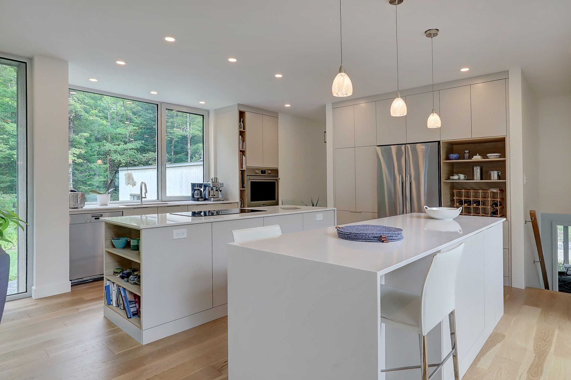 Une cuisine moderne avec des armoires blanches, deux grands &icirc;lots, un parquet clair, trois suspensions lumineuses et une vue sur l'ext&eacute;rieur.