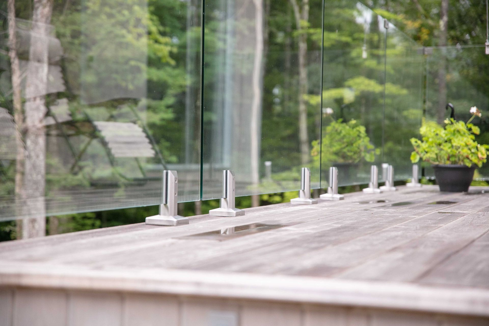 Panneaux de garde-corps en verre fix&eacute;s par des supports m&eacute;talliques argent&eacute;s sur une terrasse en bois donnant sur une for&ecirc;t.