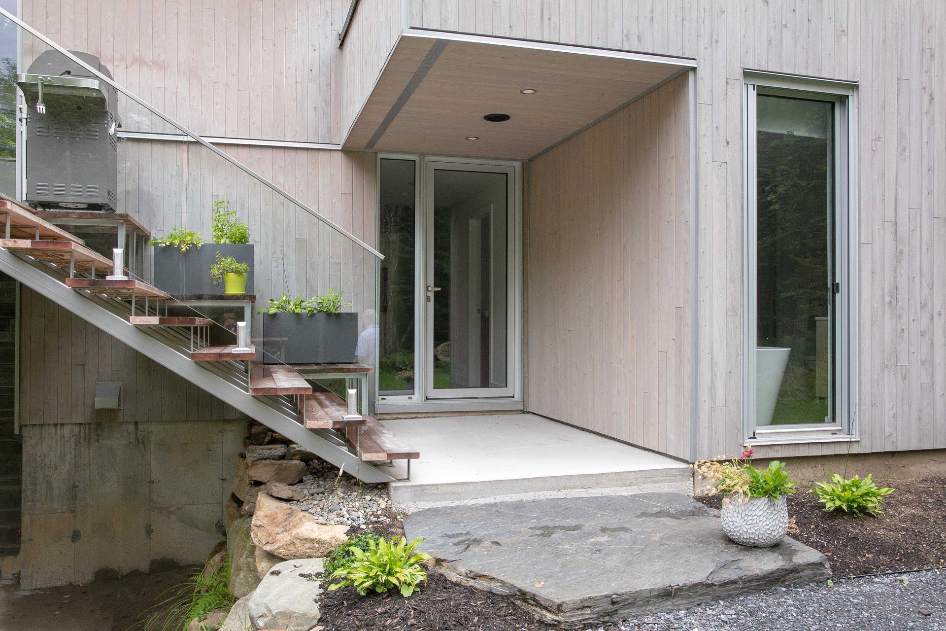 Une entr&eacute;e de maison moderne avec un bardage en bois clair, une porte vitr&eacute;e, des marches en b&eacute;ton et un escalier ext&eacute;rieur en m&eacute;tal avec des jardini&egrave;res.
