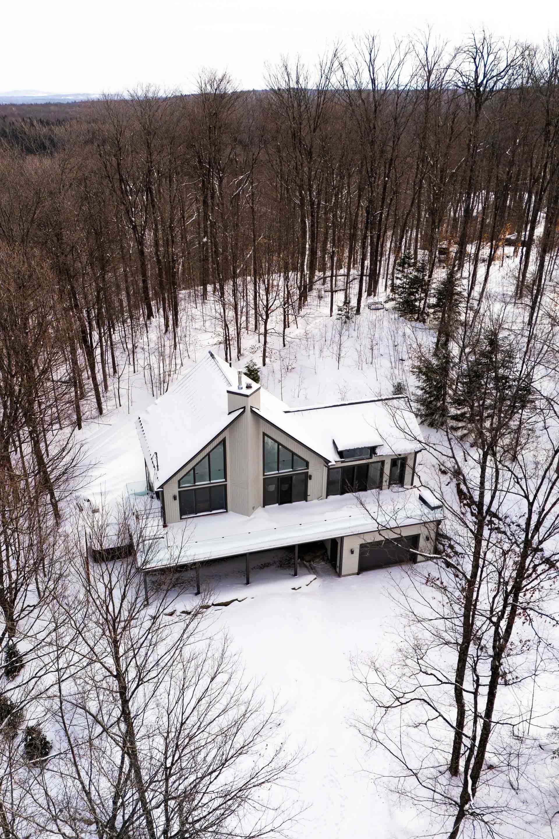 Vue a&eacute;rienne d'un chalet moderne nich&eacute; dans une for&ecirc;t enneig&eacute;e, avec des arbres d&eacute;nud&eacute;s et un toit saupoudr&eacute; de neige.