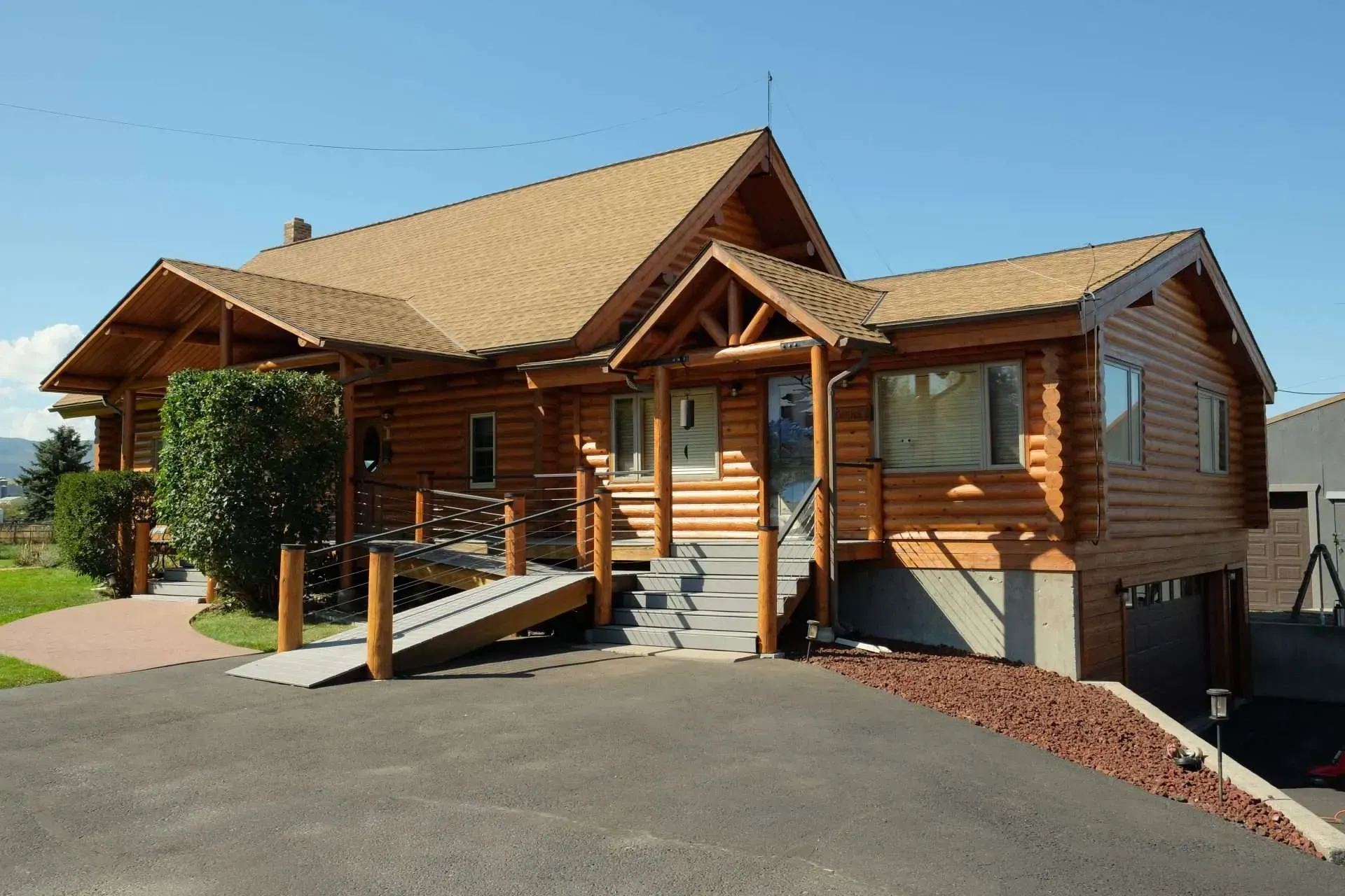 Wooden cabin with a covered porch and ramp, viewed from a paved driveway in bright daylight
