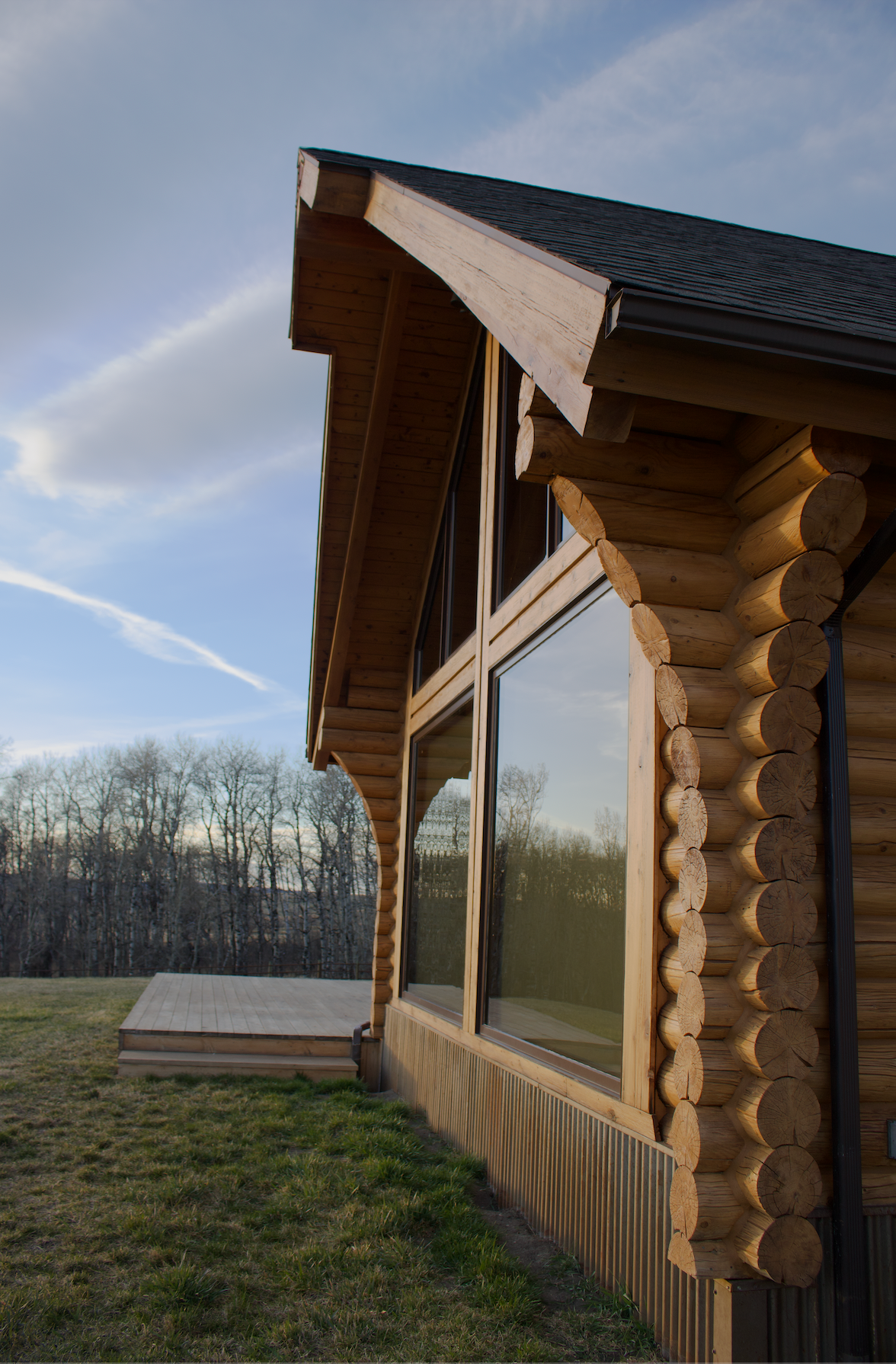 Log cabin exterior with large window, wooden deck, and grassy field under a blue sky
