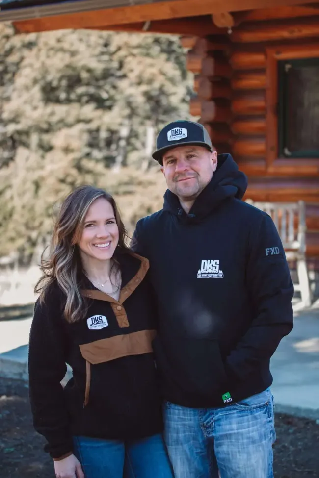 Two people in black jackets standing outside a log cabin, smiling at the camera.